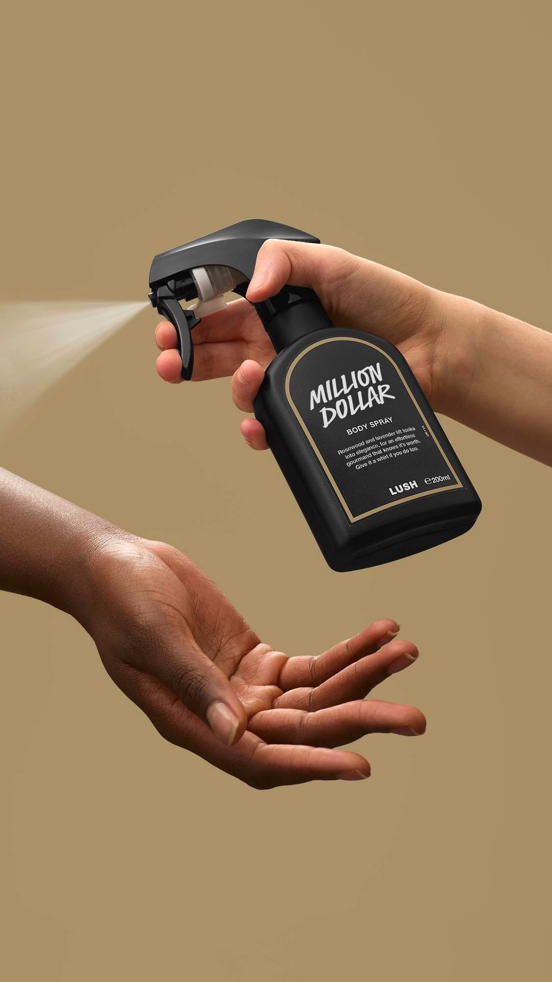 A person's hand presses the nozzle of a black Lush 'Million Dollar' body spray bottle, releasing a fine mist onto another person's open palm. The action is set against a solid, plain beige background.