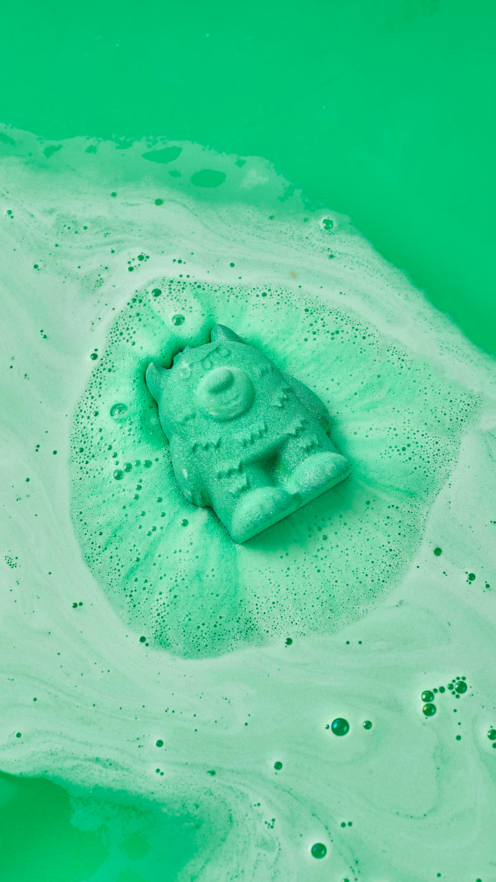 A green, three-eyed monster bath bomb fizzes and dissolves in bright green bathwater, surrounded by swirls of light green foam and bubbles.