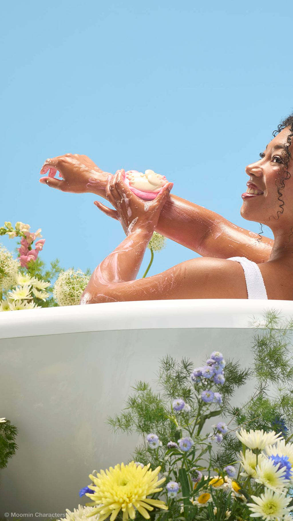 A close-up shot of a smiling person in a bathtub, lathering their arm with a pink and white "Moomin Delight" soap and body wash product. Wildflowers frame the bottom of the white tub against a clear blue sky background.
