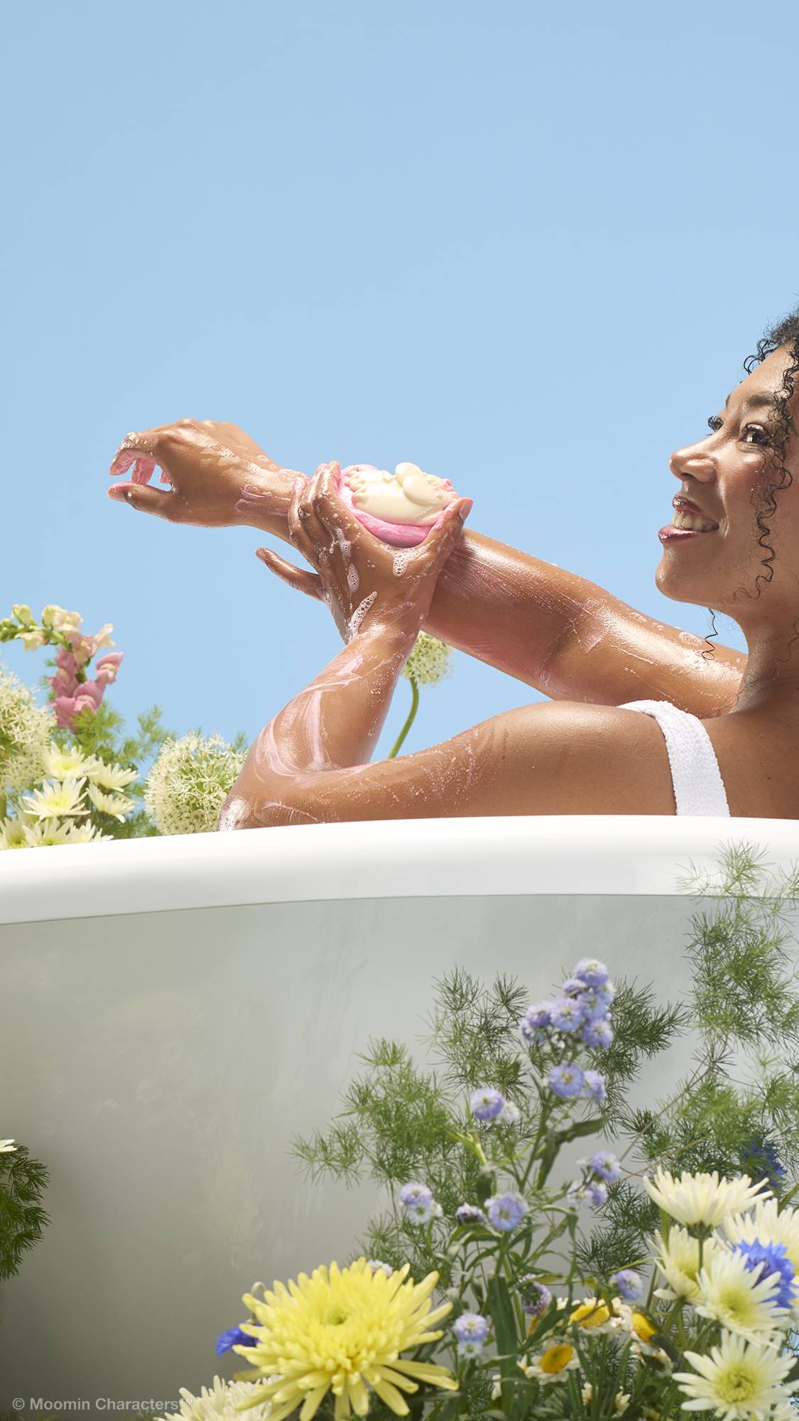 A close-up shot of a smiling person in a bathtub, lathering their arm with a pink and white "Moomin Delight" soap and body wash product. Wildflowers frame the bottom of the white tub against a clear blue sky background.