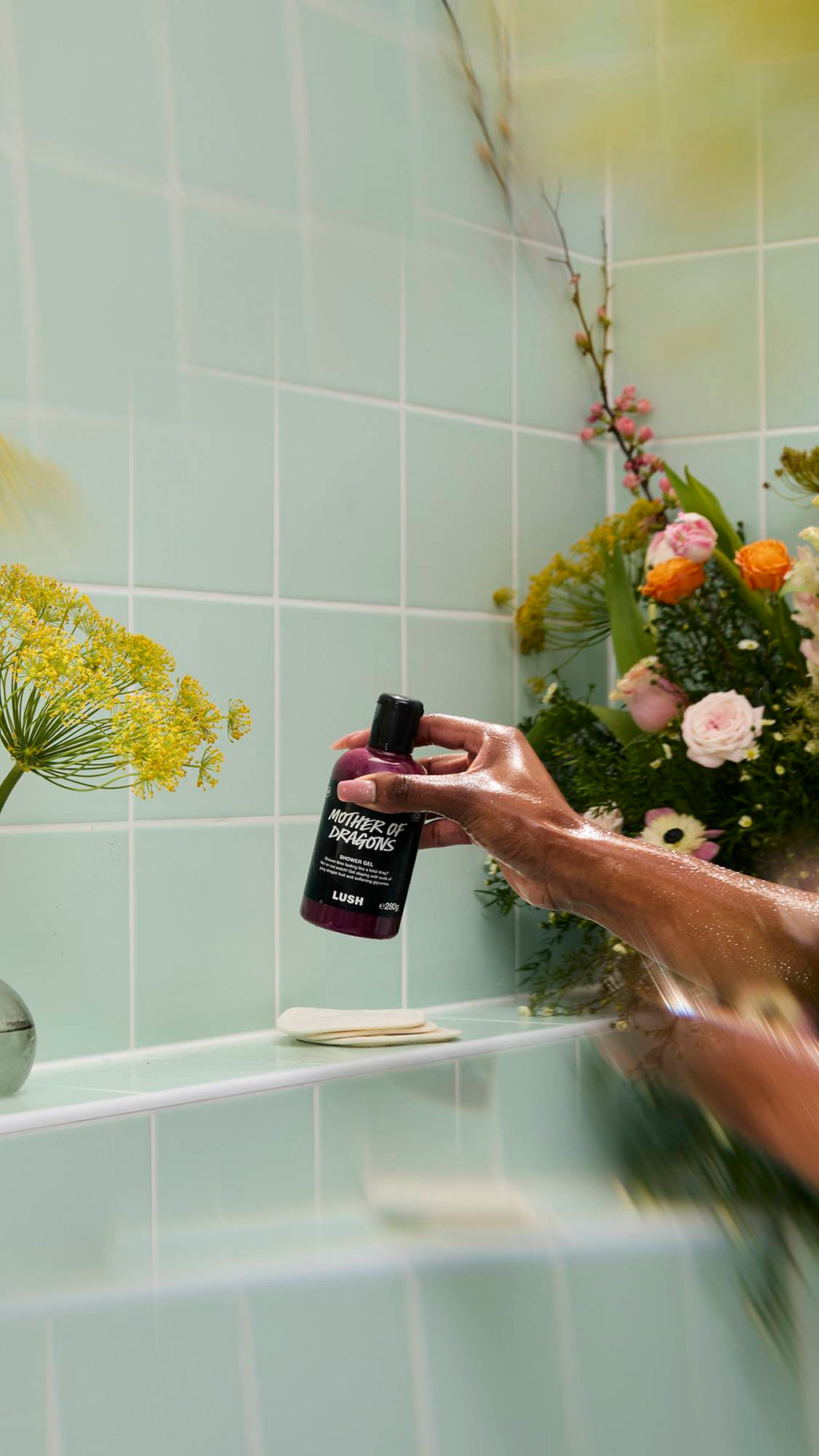 A bottle of purple, shimmery "Mother of Dragons" Shower Gel sits on a white tiled shelf. The label mentions "juicy dragon fruit and softening glycerine." Bubbles and greenery surround the bottle against a mint green tiled background.