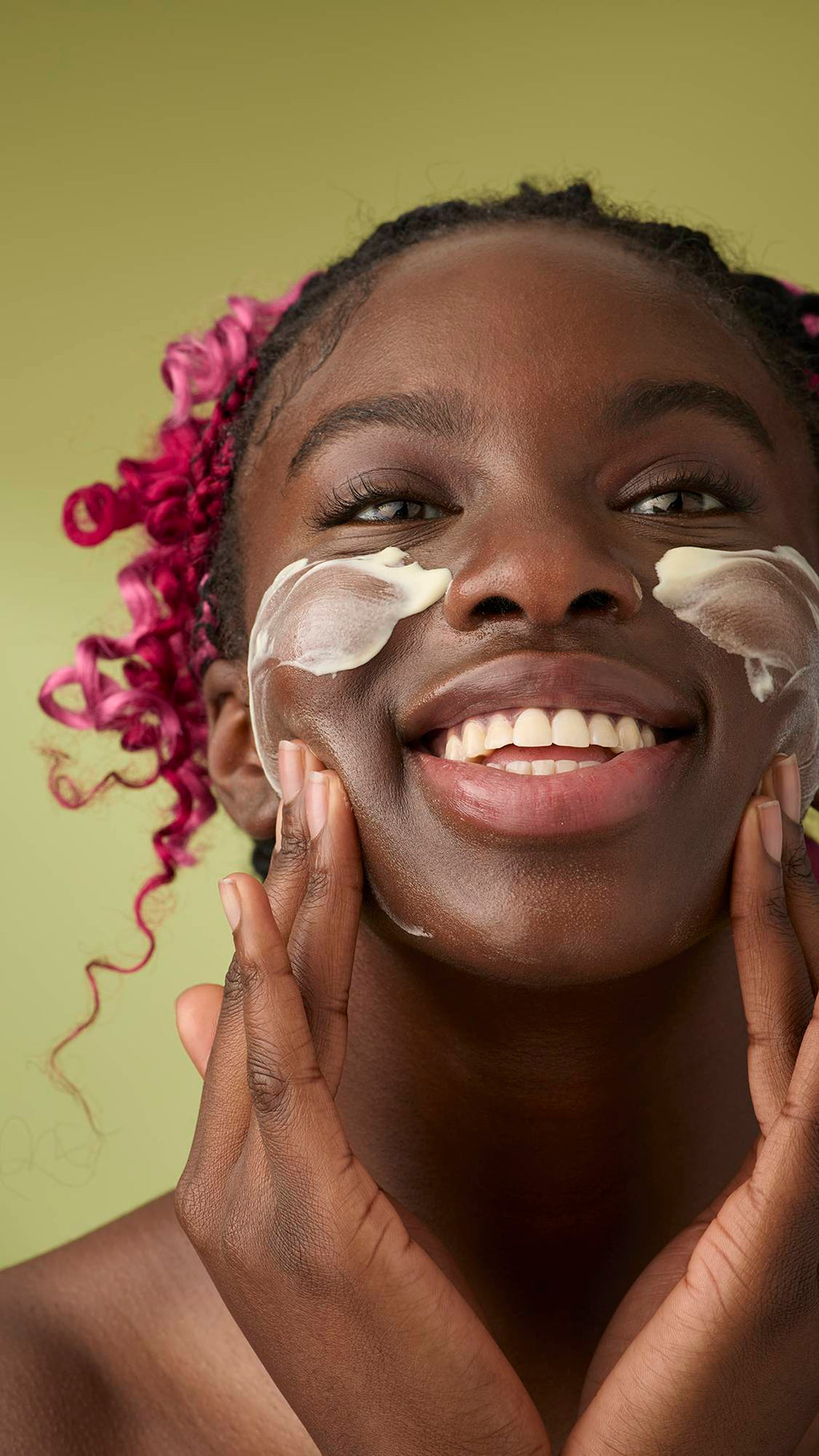 A woman with pink and black braids smiles as she applies a smooth, ivory Oat facial cleanser to her cheeks. The thick, creamy product stands out against her skin and the solid olive-green background, emphasising its rich texture.