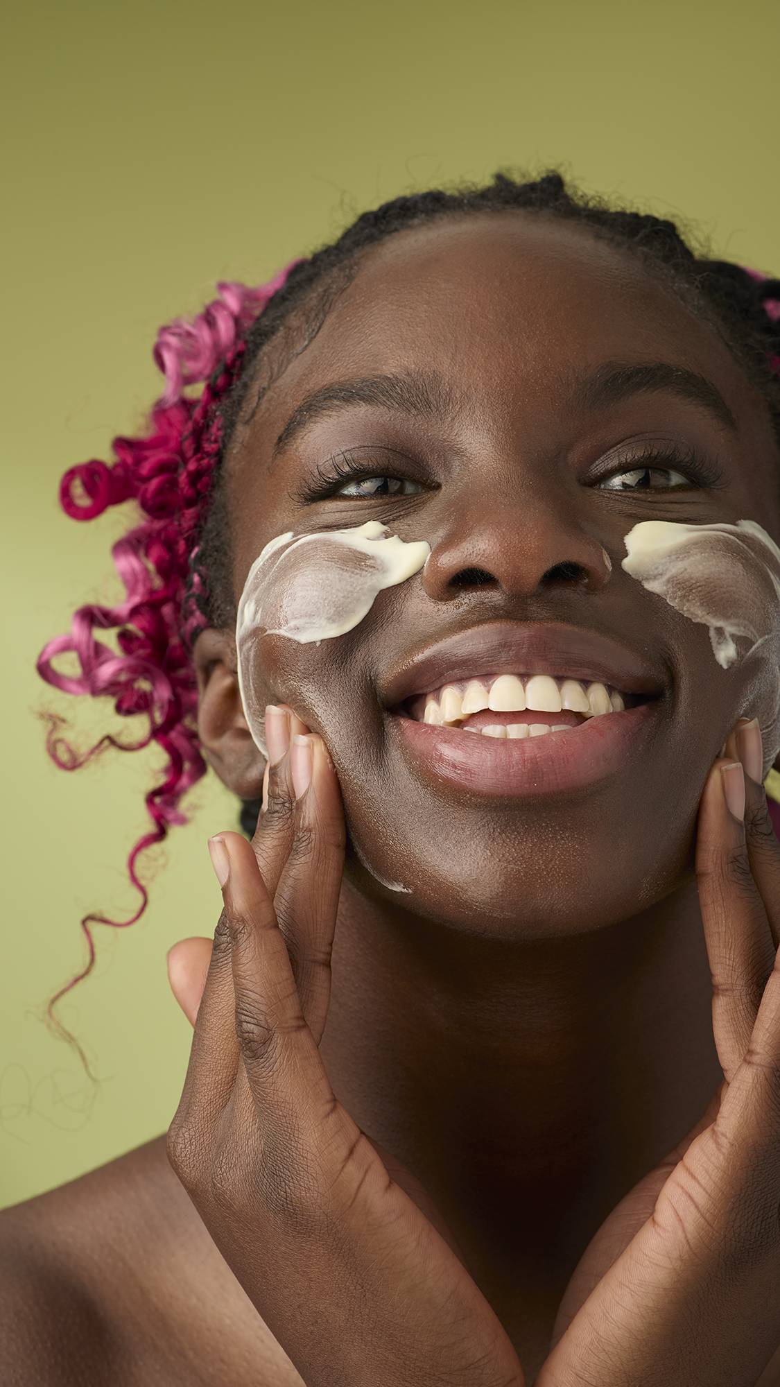 A woman with pink and black braids smiles as she applies a smooth, ivory Oat facial cleanser to her cheeks. The thick, creamy product stands out against her skin and the solid olive-green background, emphasising its rich texture.