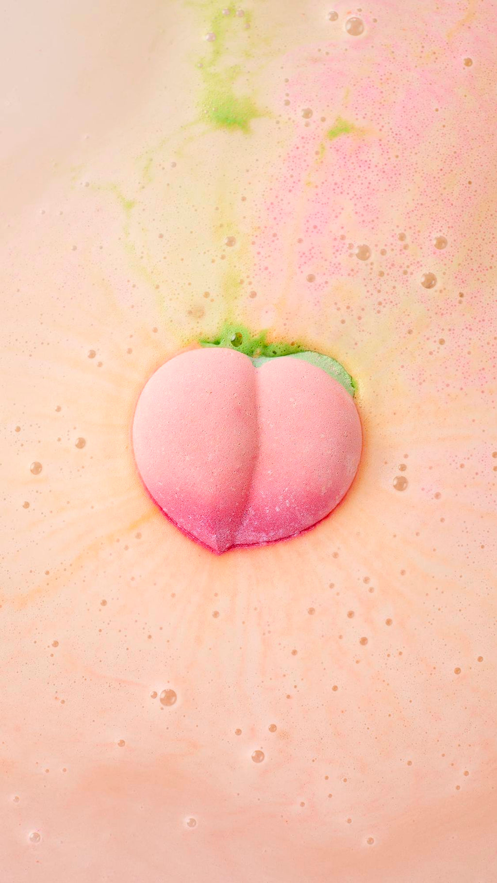 A peach-shaped bath bomb fizzes in the centre of a tub, releasing swirls of vibrant pink and lime green into creamy, peach-coloured water. The surface is covered in a delicate layer of bubbles and frothy foam.