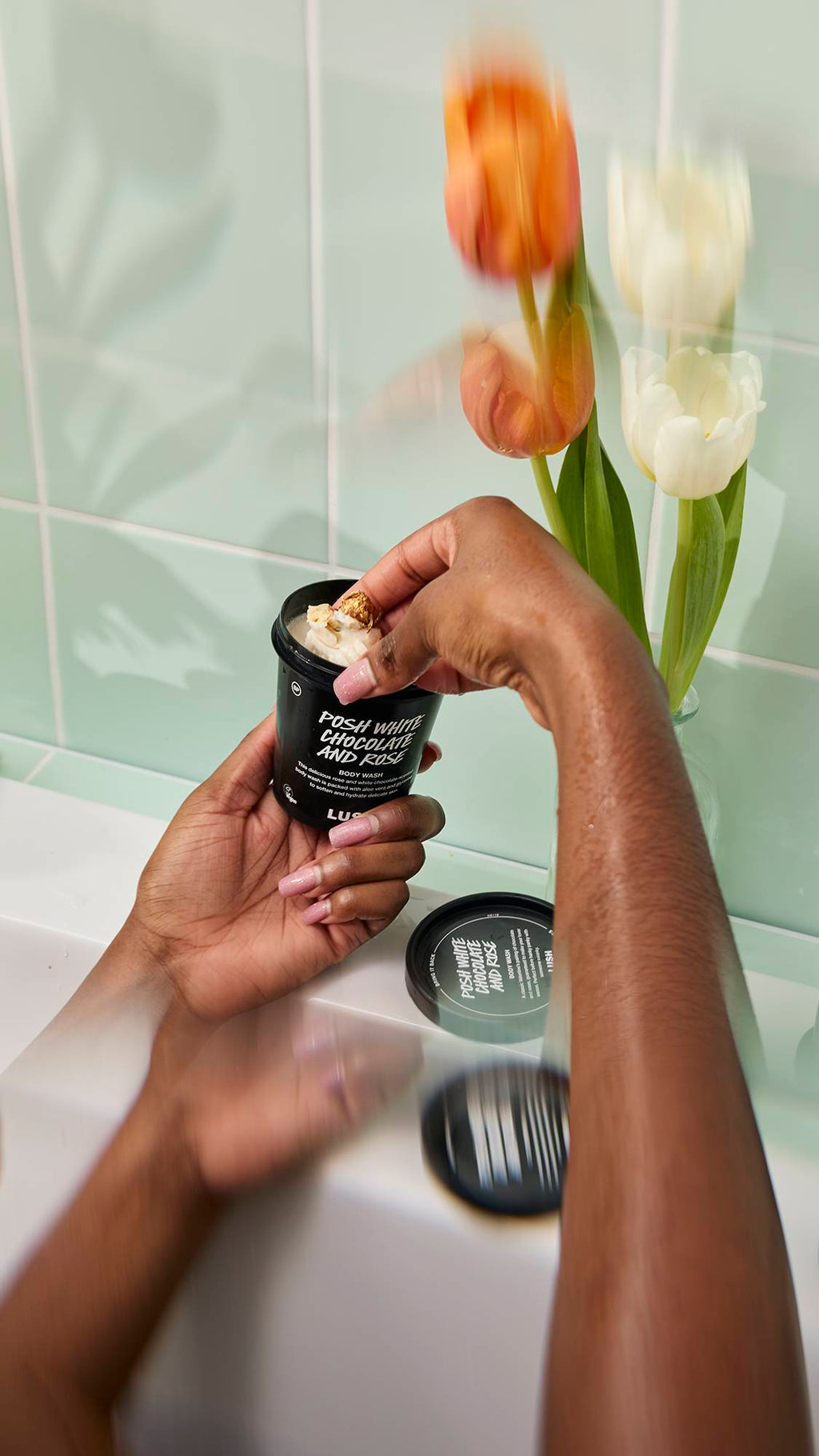 A person sitting in a bath scoops thick, white "Posh White Chocolate and Rose" Body Wash from a black pot. The scene features wet hands and blurred tulips against a mint green tiled background.