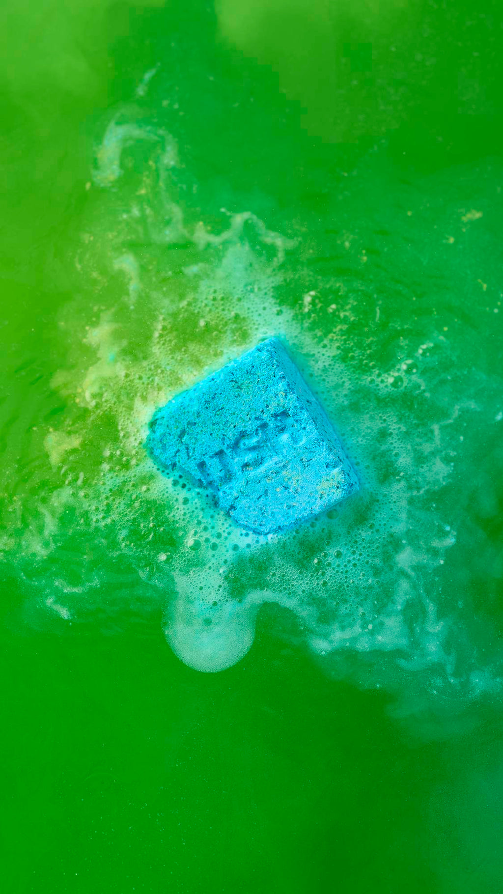 The blue and yellow, cubed "Salt Water Soother" bath bomb, with "LUSH" debossed on its surface, dissolves in bright green bathwater. It is surrounded by a patch of white, frothy bubbles as the vibrant blue and green colours begin to swirl together.