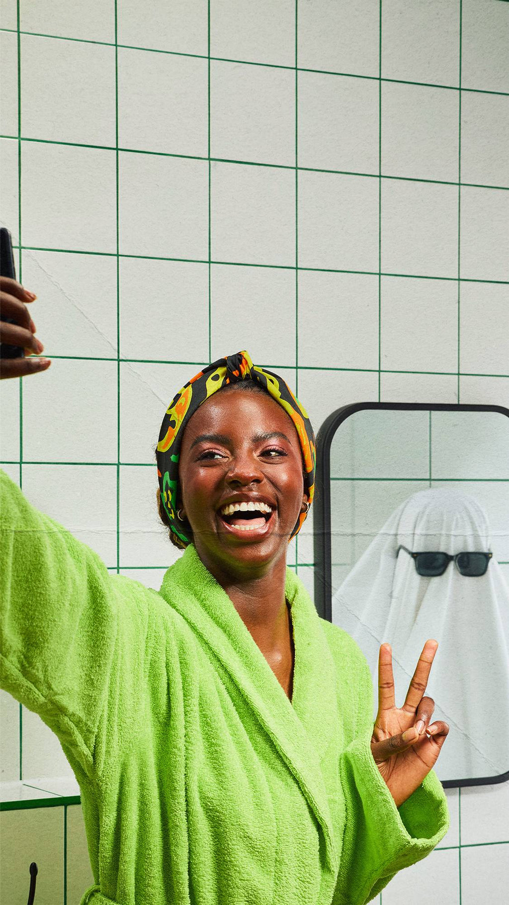 A model in a lime green dressing gown and patterned headscarf, fashioned from a knot wrap, takes a selfie in a bathroom. Their reflection in the mirror shows a person dressed as a ghost under a white sheet, wearing sunglasses with a rubber duck.