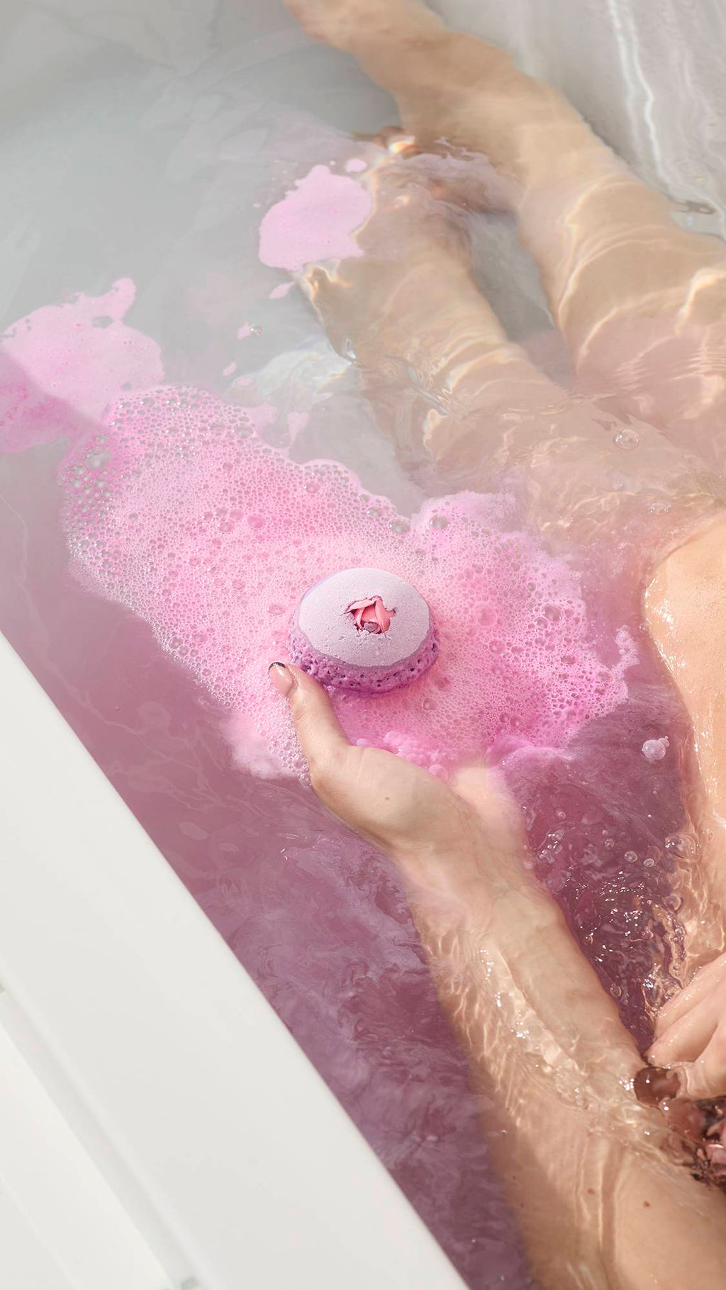 A hand holds the Sex Bomb Bath Bomb over a bathtub of frothy pink water while a person relaxes in the background.