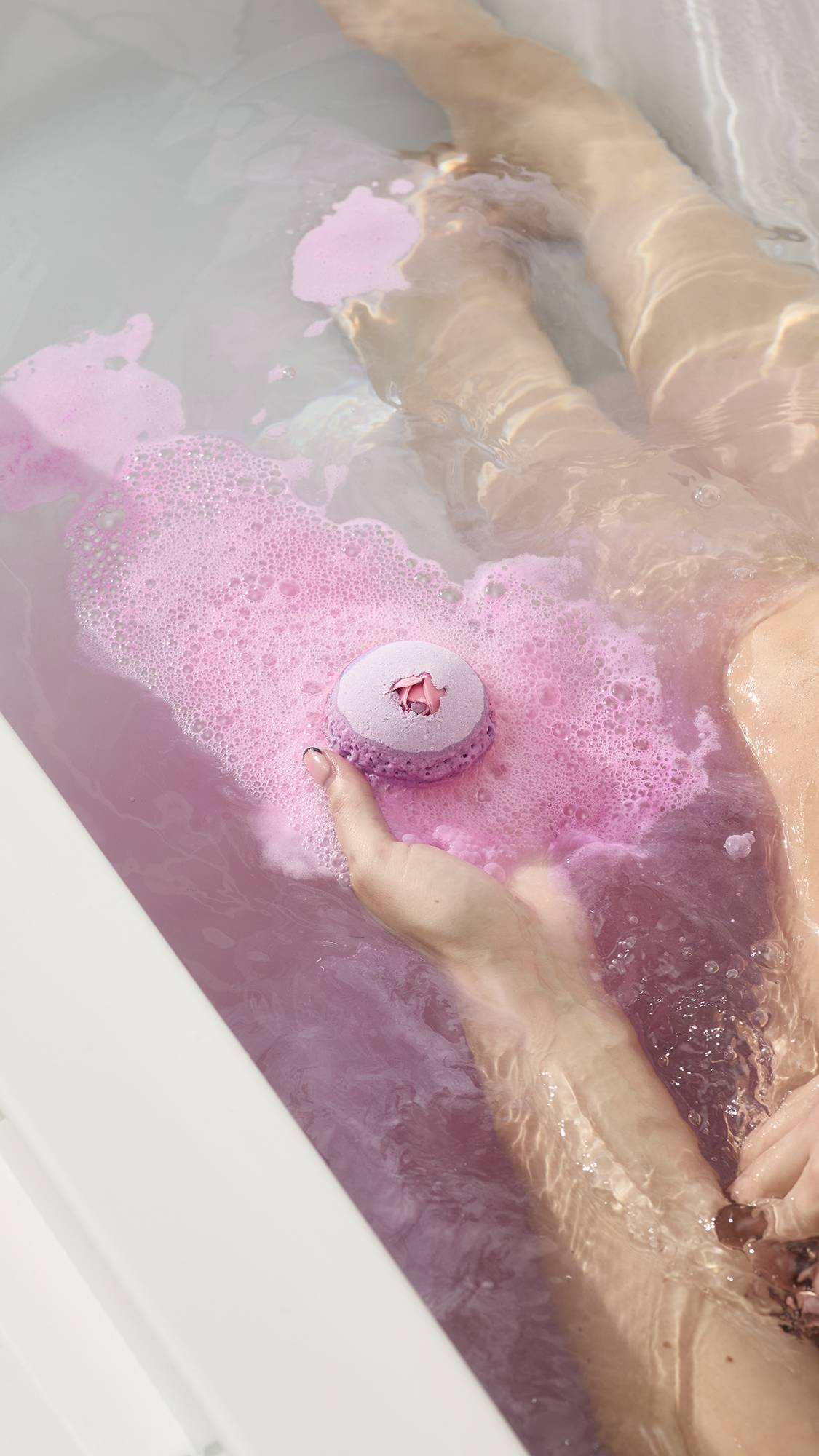 A hand holds the Sex Bomb Bath Bomb over a bathtub of frothy pink water while a person relaxes in the background.