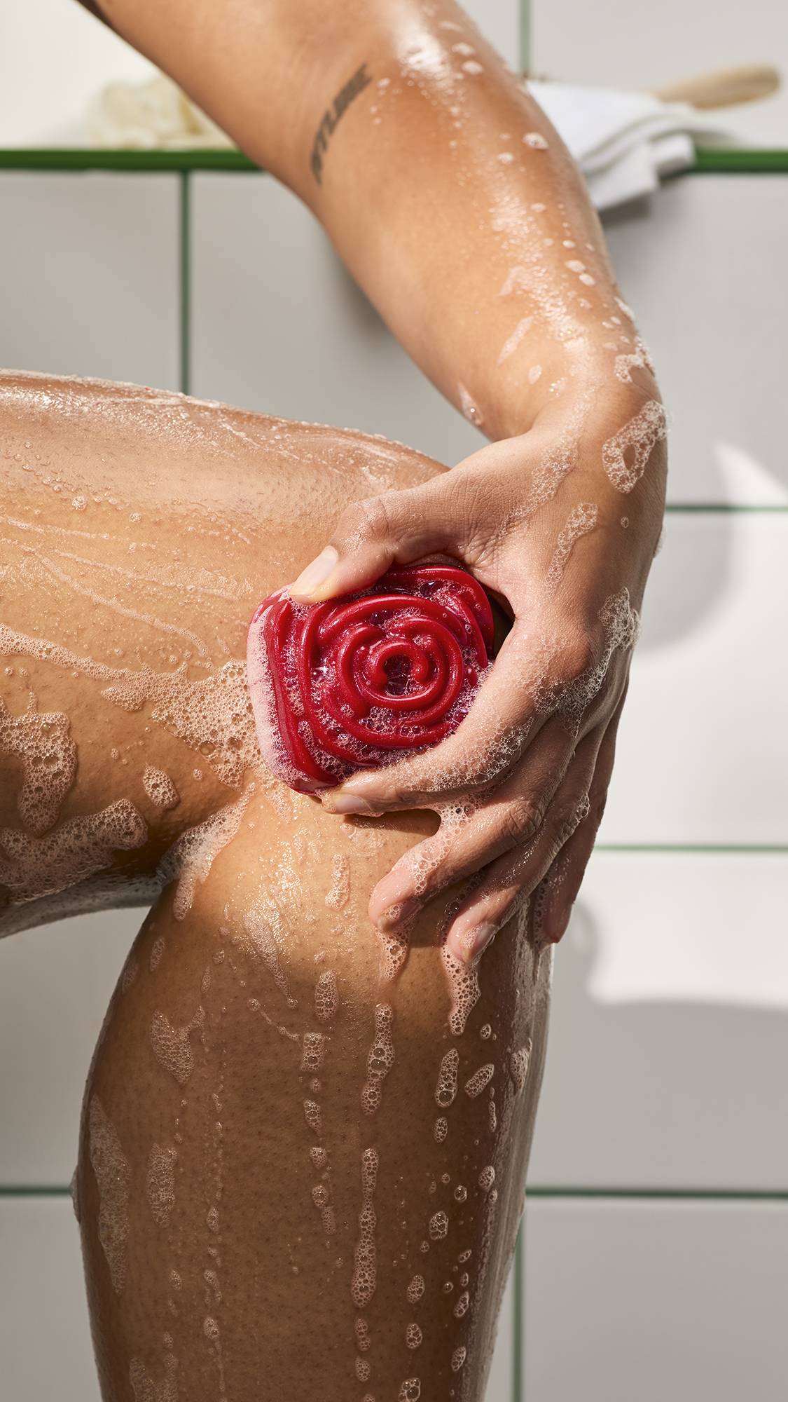 A hand lathers a wet leg with a red, rose-shaped shower gummy soap. Bubbles and foam run down the skin against white bathroom tiles with green grout.