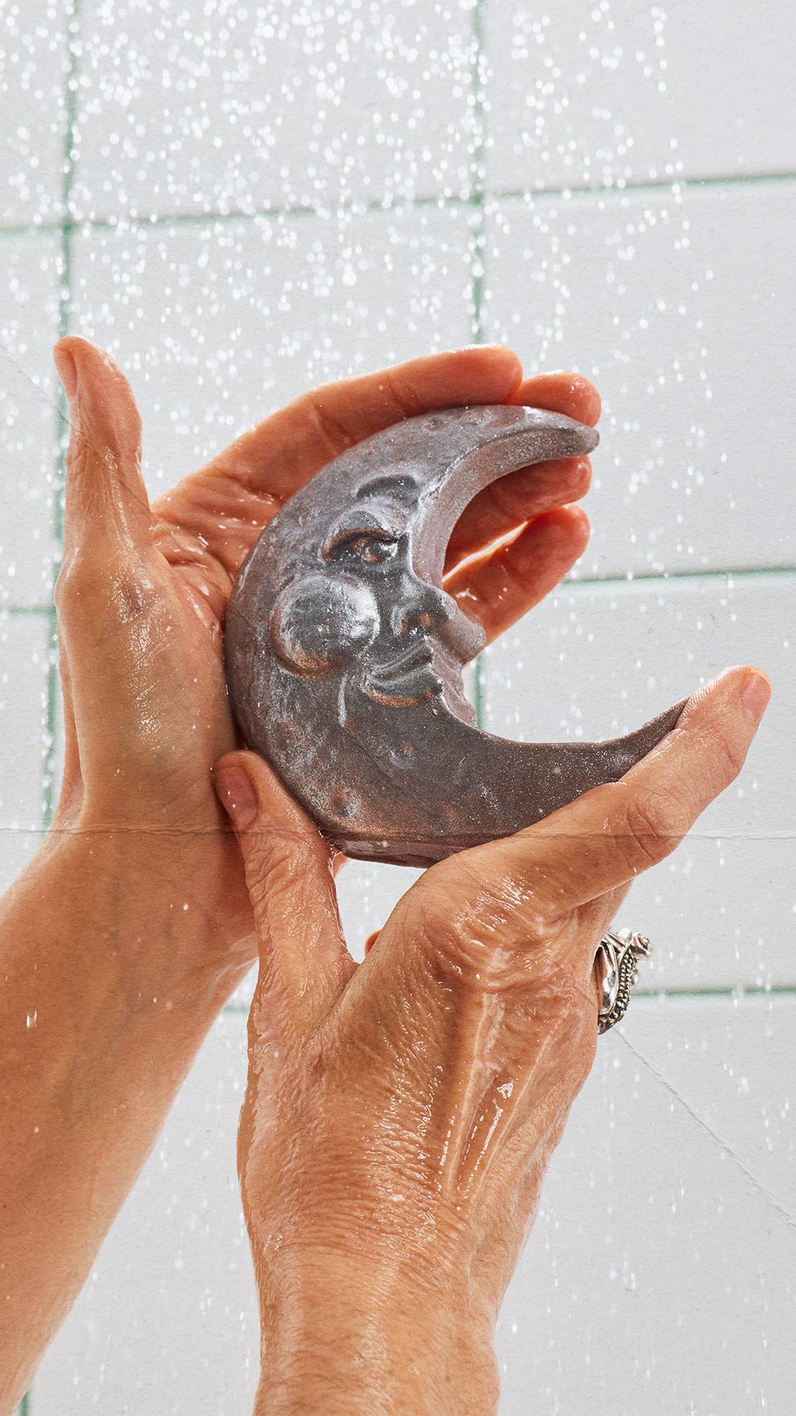 Two wet hands cupping a silver, crescent moon-shaped soap bar featuring a stylised face. Water spray falls around the hands against a background of white tiles with green grout lines.