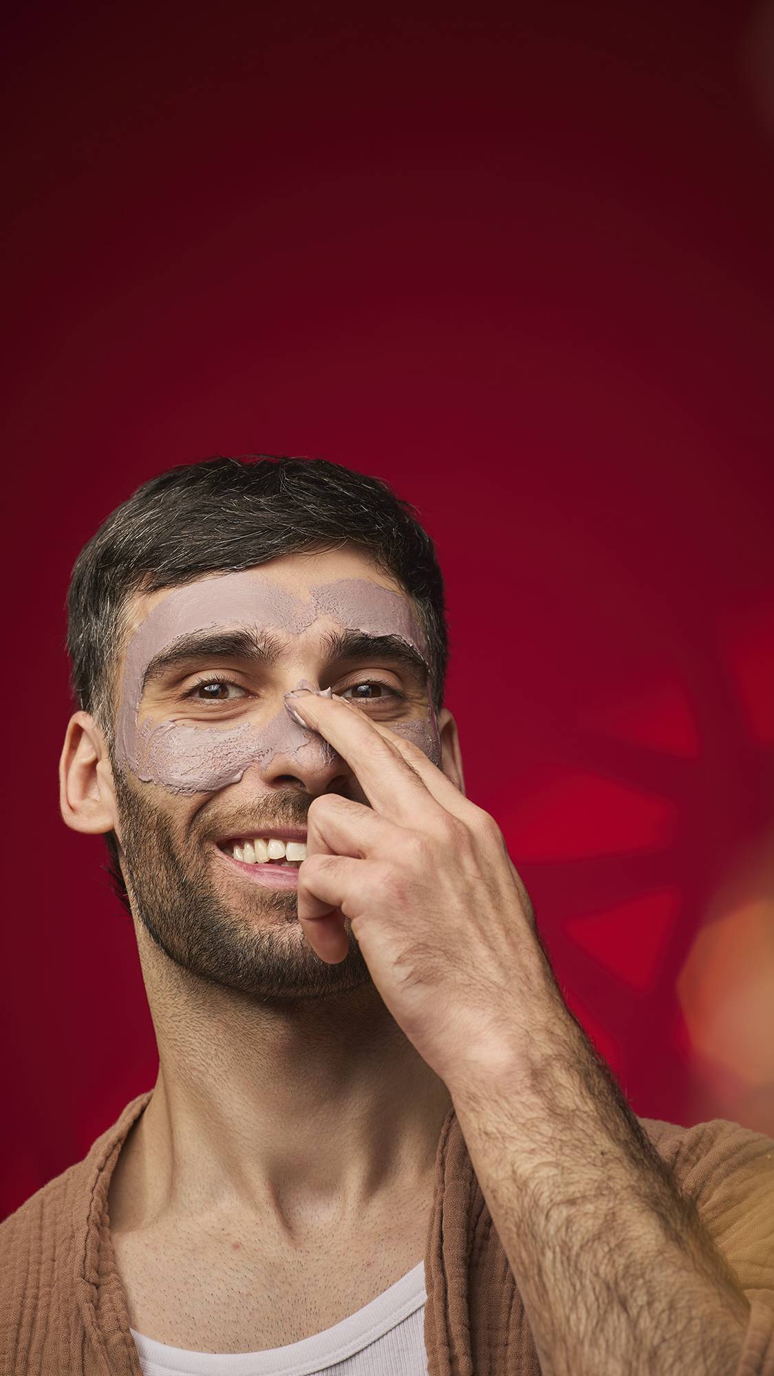 A person smiles while applying a thick, mauve-brown Sipak face mask to their nose and cheek area. They are wearing a brown robe against a deep red background with decorative bokeh patterns.