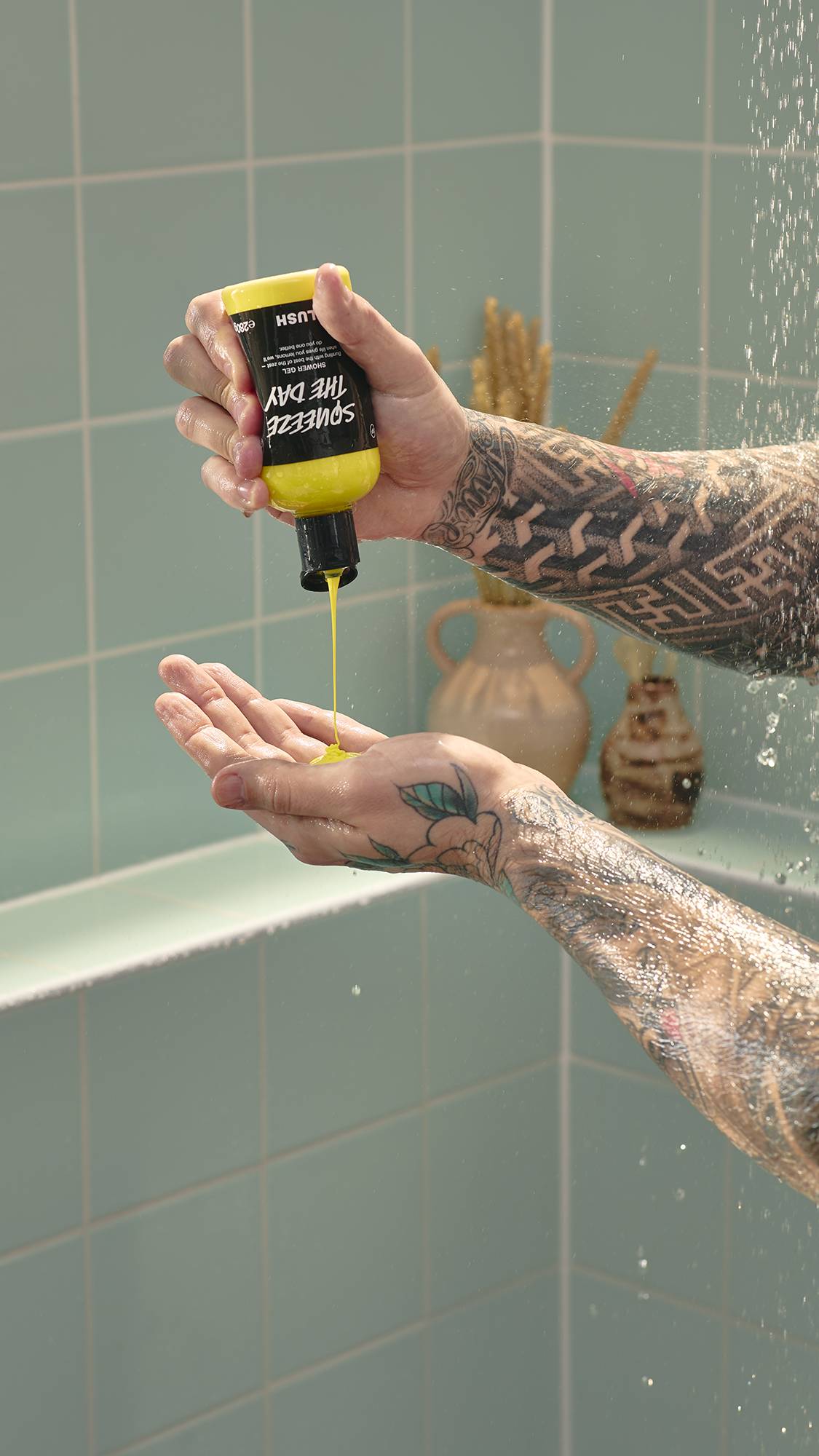 A model with heavily tattooed arms pours bright yellow "Squeeze The Day" shower gel from a Lush bottle into their open palm. They are standing in a shower with pale green tiles and a decorative vase in the background.