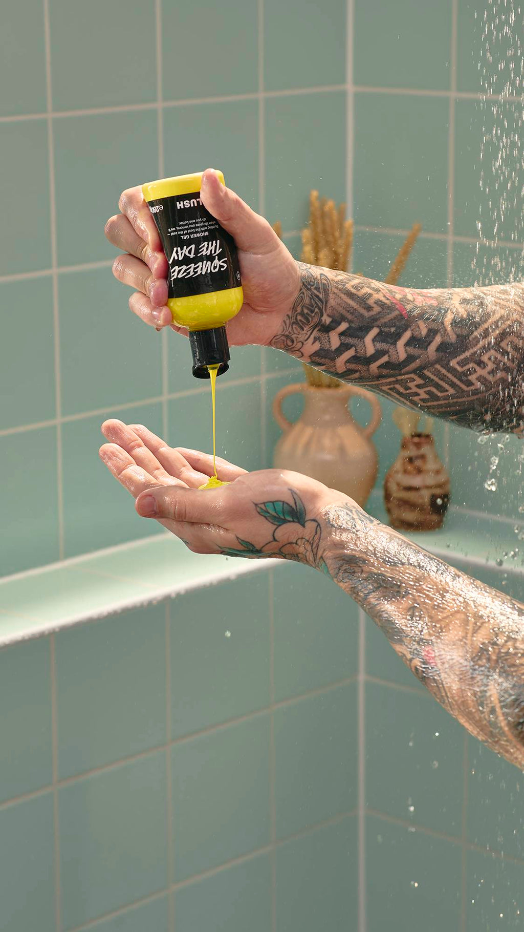 A model with heavily tattooed arms pours bright yellow "Squeeze The Day" shower gel from a Lush bottle into their open palm. They are standing in a shower with pale green tiles and a decorative vase in the background.