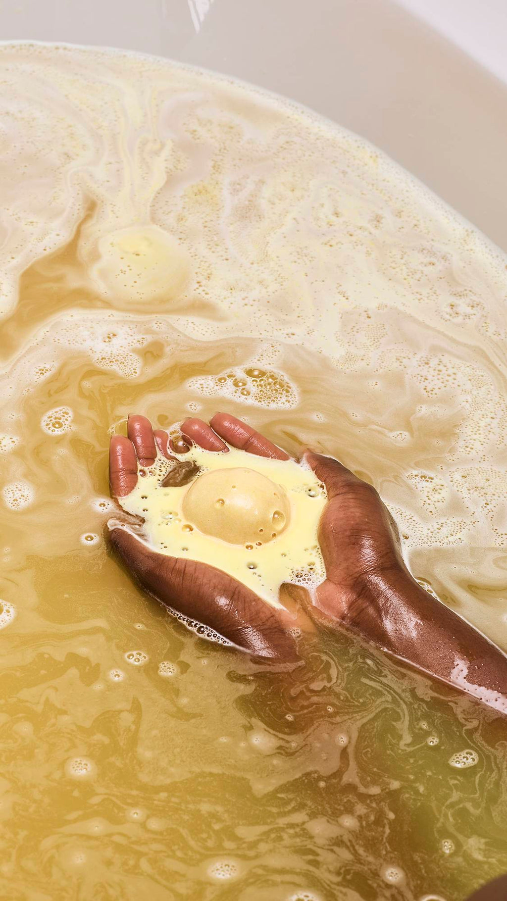 A model with deep-toned skin holds a shimmering gold "Sticky Dates" bath bomb in their cupped hands as it dissolves. It releases thick, creamy yellow foam that swirls into the golden, bubbling bathwater