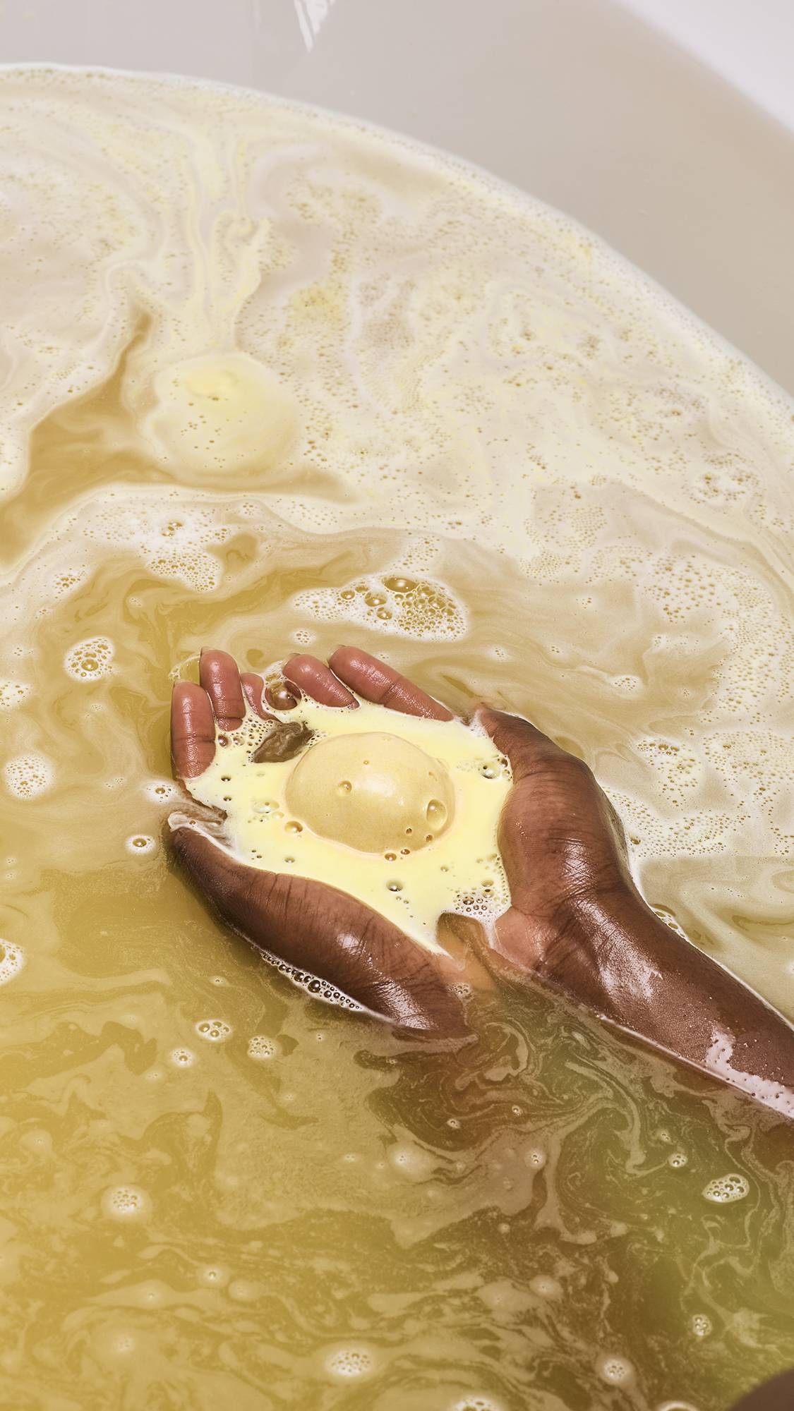 A model with deep-toned skin holds a shimmering gold "Sticky Dates" bath bomb in their cupped hands as it dissolves. It releases thick, creamy yellow foam that swirls into the golden, bubbling bathwater