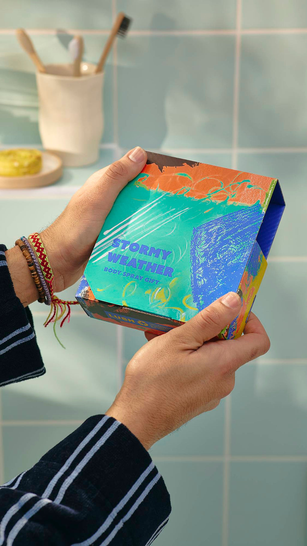 A model wearing a striped dressing gown holds a Lush ‘Stormy Weather’ gift box. The square box features a vibrant, abstract design in teal, orange, and blue. In the background, toothbrushes sit in a cup against blue bathroom tiles.