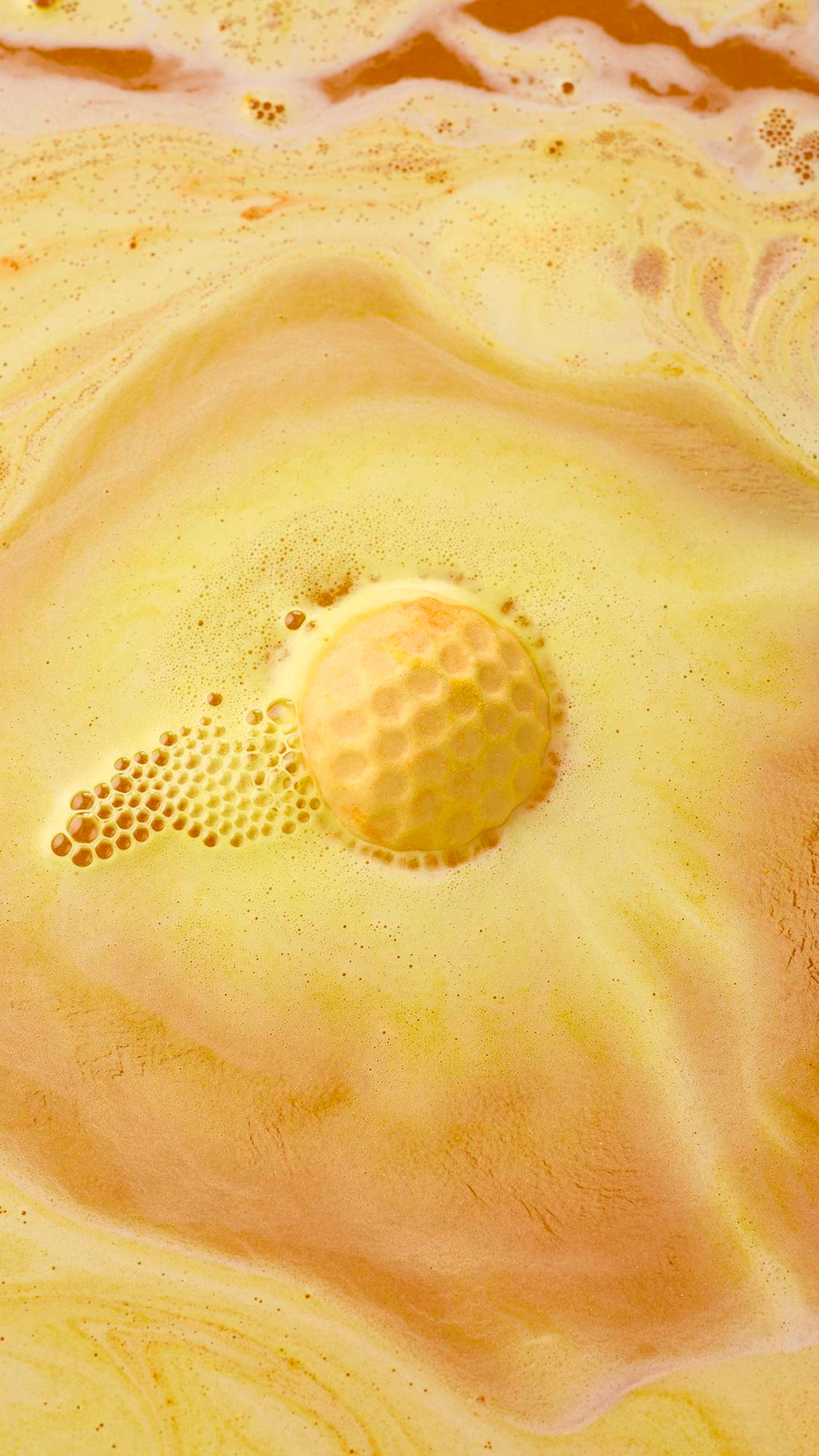An aerial view of a golden-yellow, "Sweet As Honey" bath bomb dissolving in water. It is surrounded by frothy bubbles and vibrant swirls of amber and yellow, creating a creamy, marbled effect across the surface.