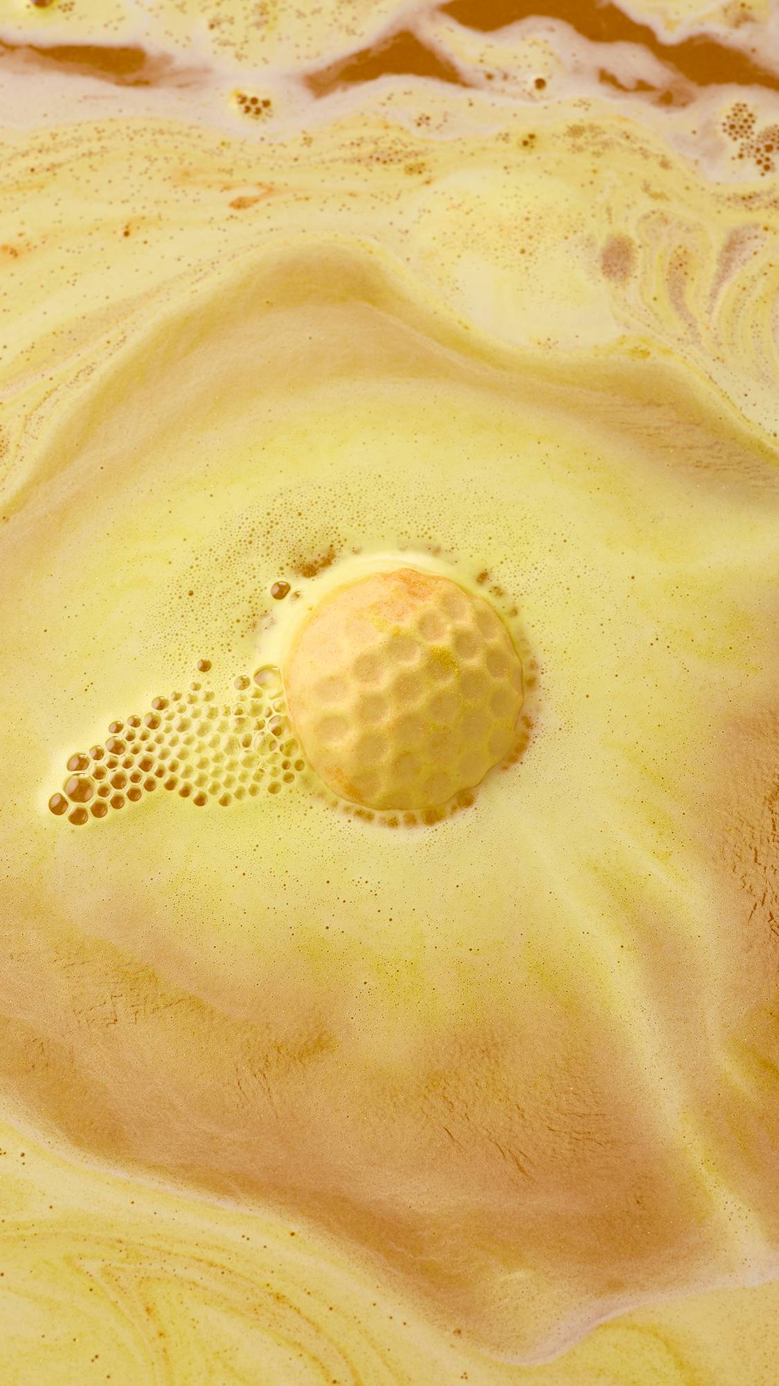 An aerial view of a golden-yellow, "Sweet As Honey" bath bomb dissolving in water. It is surrounded by frothy bubbles and vibrant swirls of amber and yellow, creating a creamy, marbled effect across the surface.