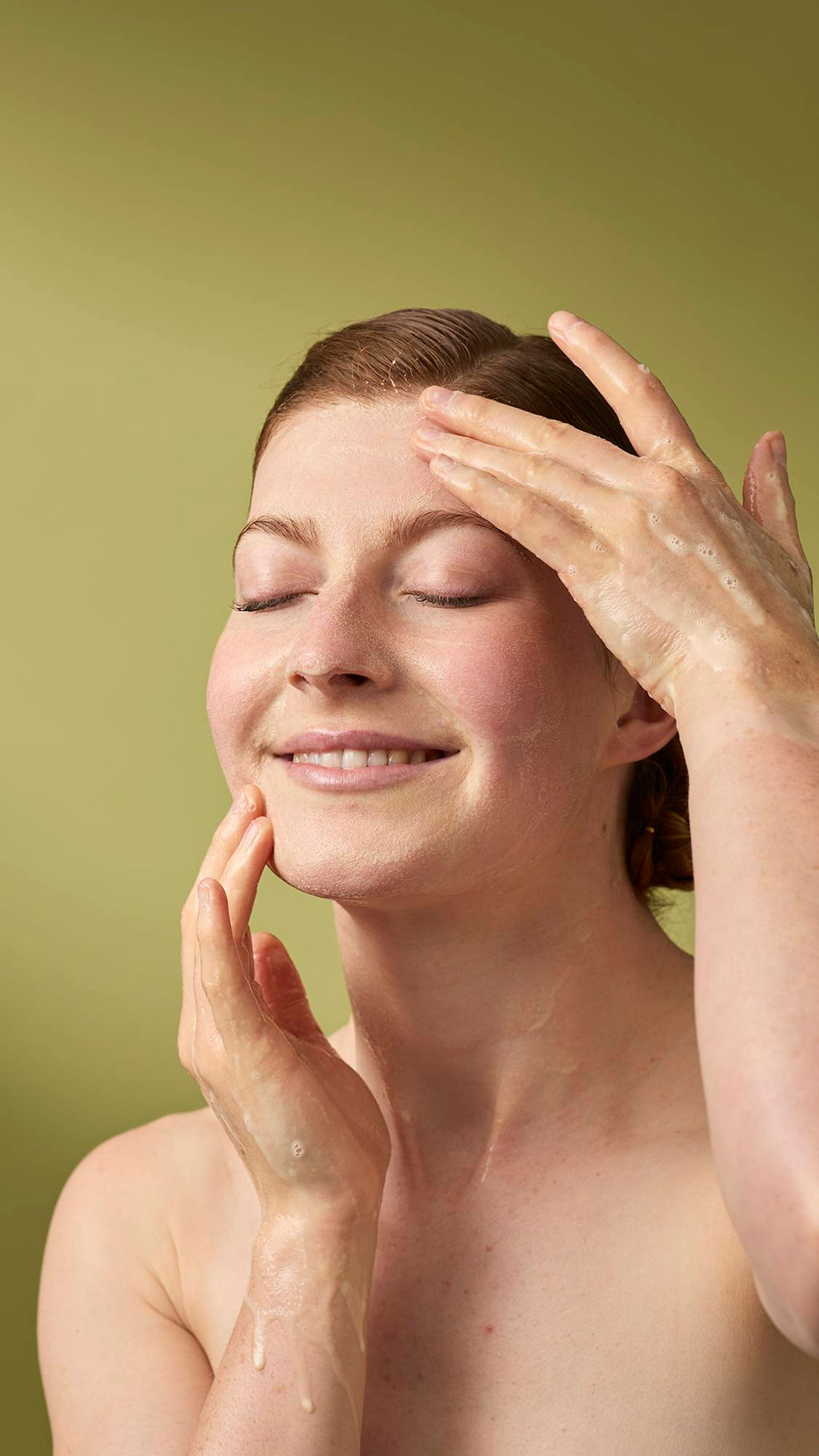 A person with their eyes closed and a gentle smile massages a foamy cleanser into their face. Their wet hands are covered in light soap suds, and they stand against a solid olive-green background.