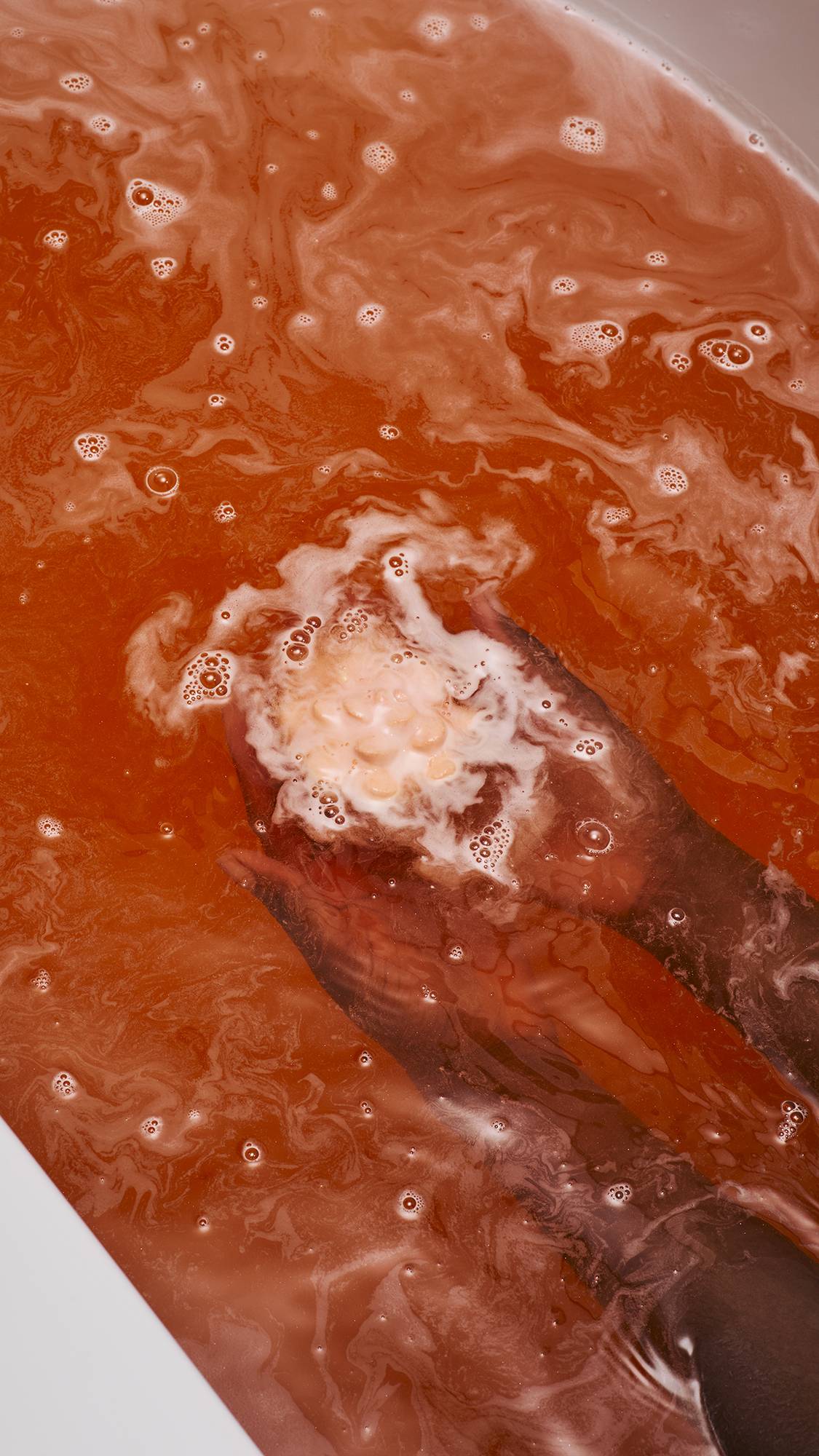 A vibrant coral-coloured "Comforter Rose" bath bomb held by two hands just beneath the surface of orange, foamy bathwater. The bath bomb is partially covered in thick white bubbles, which swirl and spread across the warm-toned water.