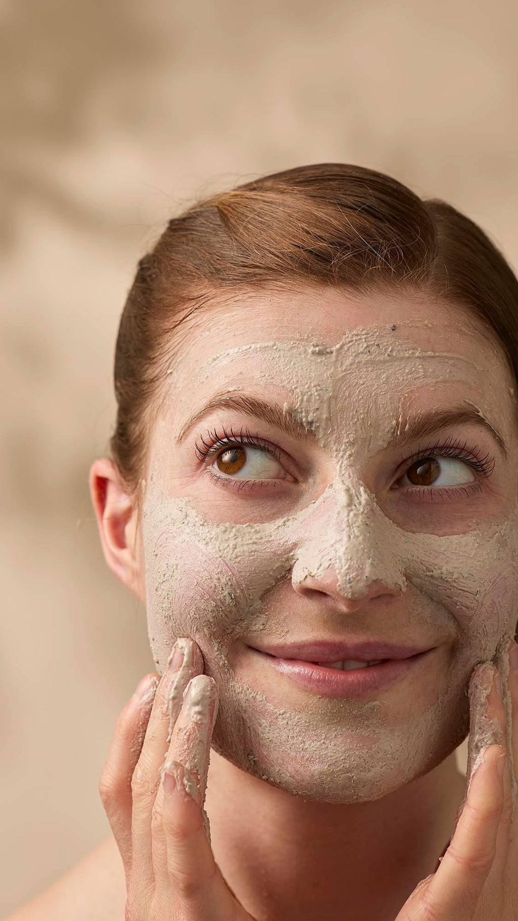 A close-up of a smiling model with reddish-brown hair tied back, applying a textured, beige face mask. They look off-camera with a playful expression while using their fingers to blend the thick cleanser across their face, avoiding the eyes.