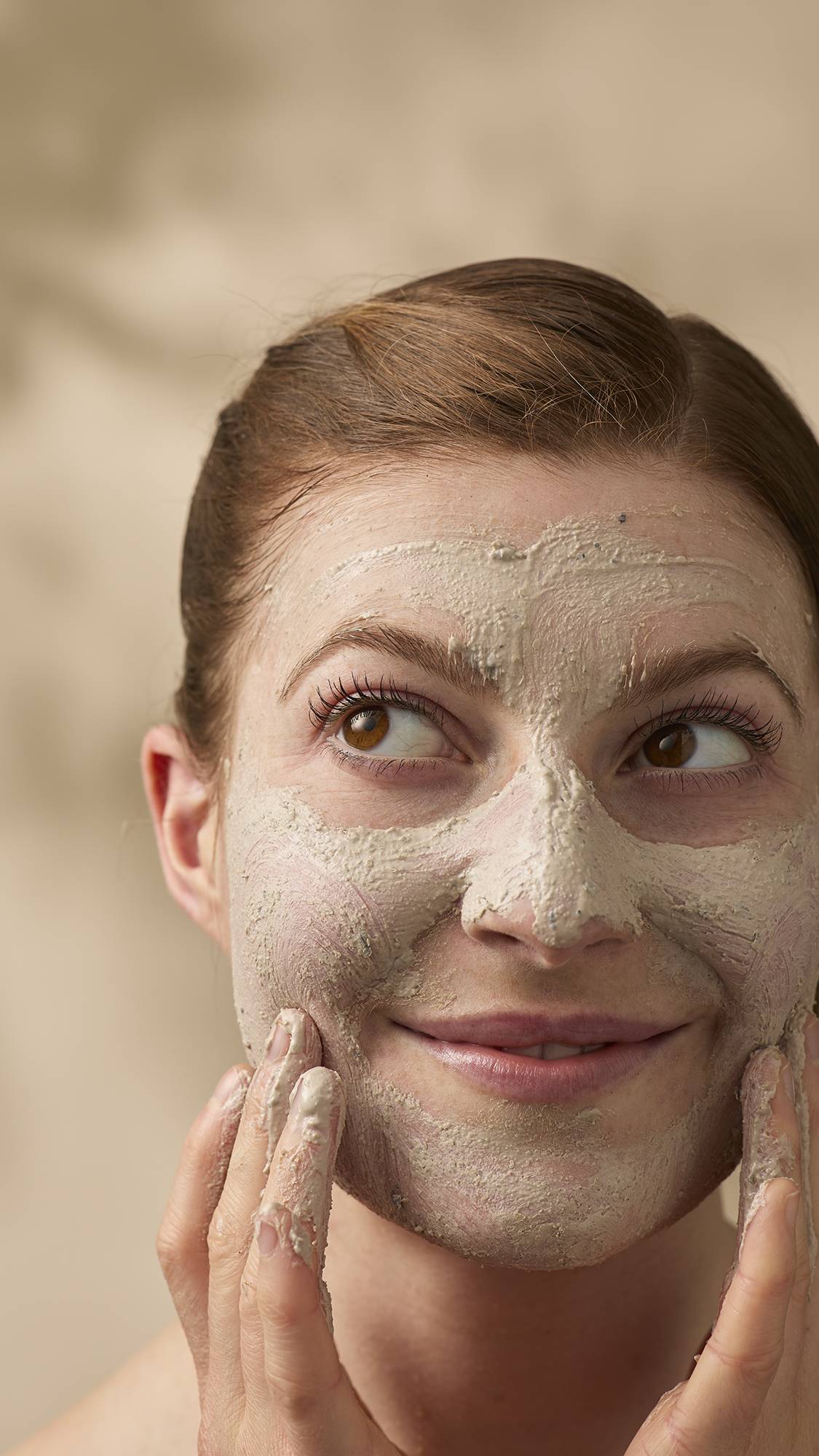 A close-up of a smiling model with reddish-brown hair tied back, applying a textured, beige face mask. They look off-camera with a playful expression while using their fingers to blend the thick cleanser across their face, avoiding the eyes.