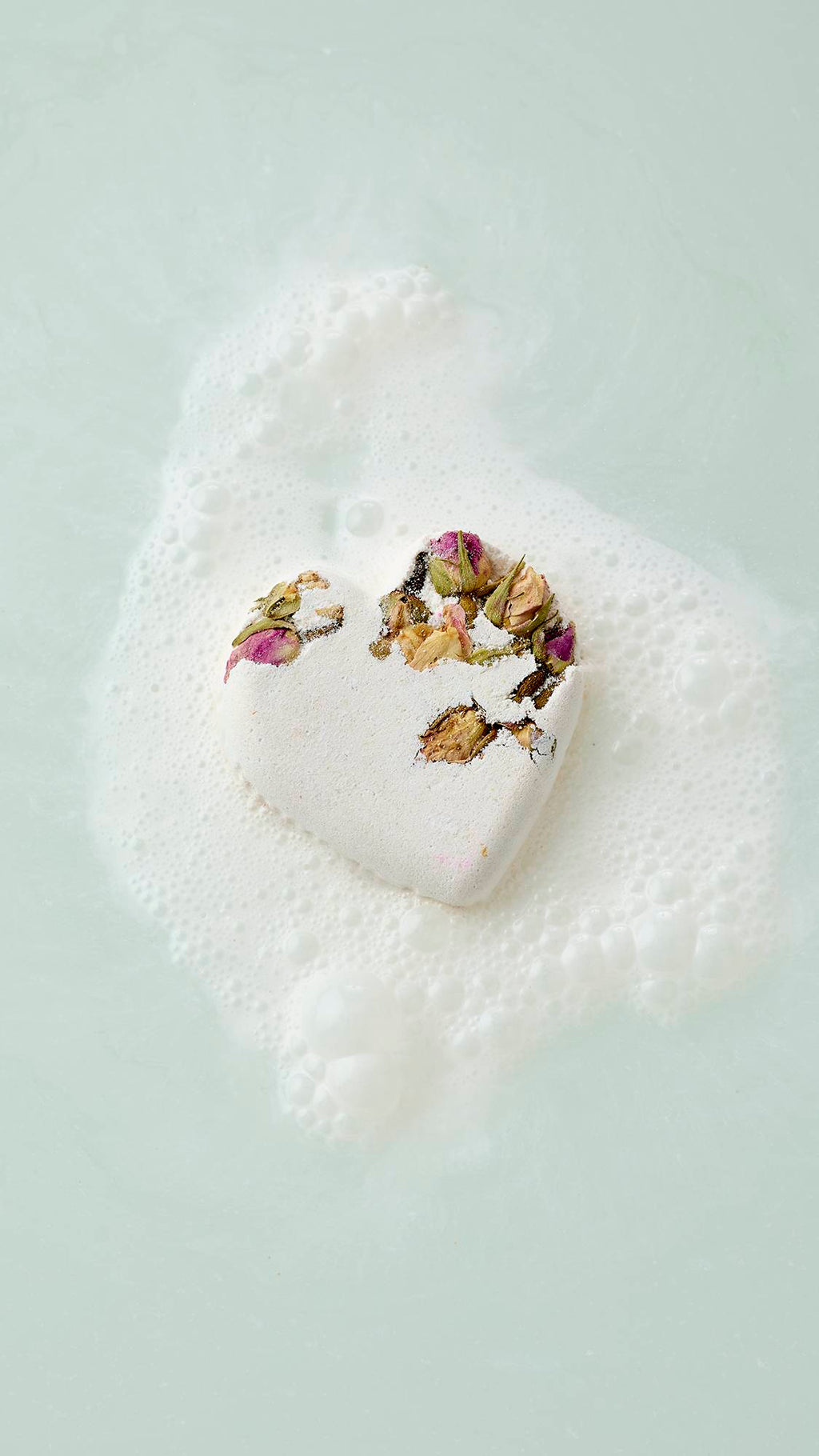 The image shows an aerial view of a white, heart-shaped Tisty Tosty bath bomb decorated with dried rosebuds floating in pale water. It is surrounded by a patch of white, frothy bubbles as it begins to dissolve.