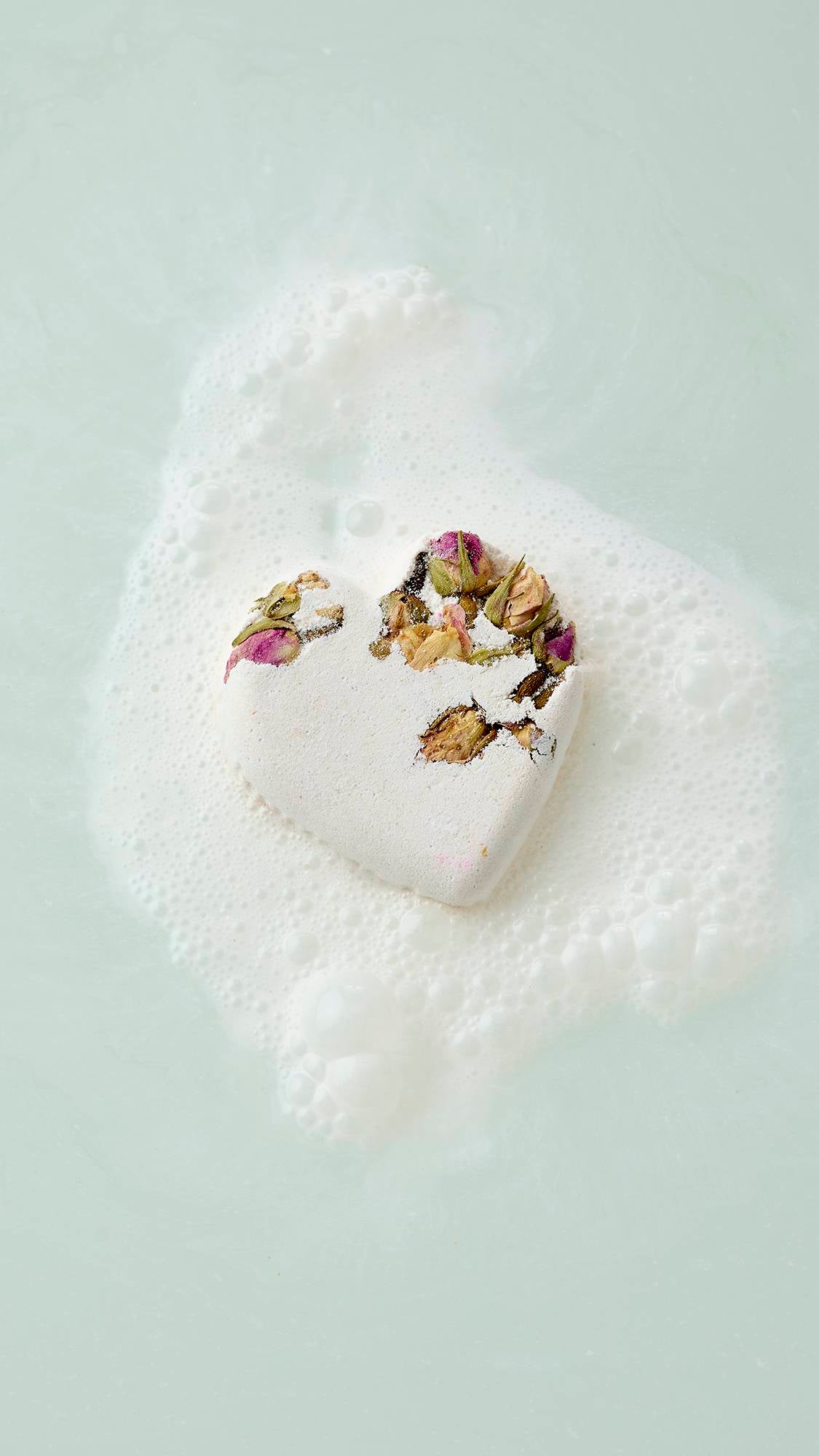 The image shows an aerial view of a white, heart-shaped Tisty Tosty bath bomb decorated with dried rosebuds floating in pale water. It is surrounded by a patch of white, frothy bubbles as it begins to dissolve.