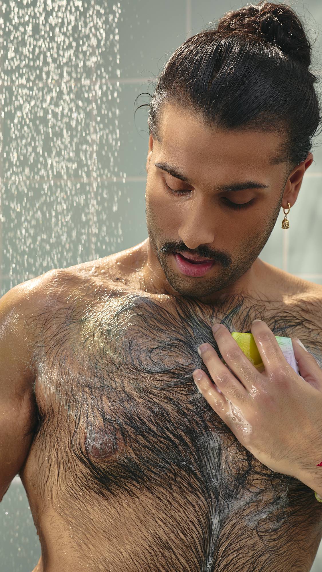 The image shows a model with dark hair in a bun. There is a running shower behind on pale green tiles. The model is using the "To The Moon and Back" soap slice to lather up their chest.