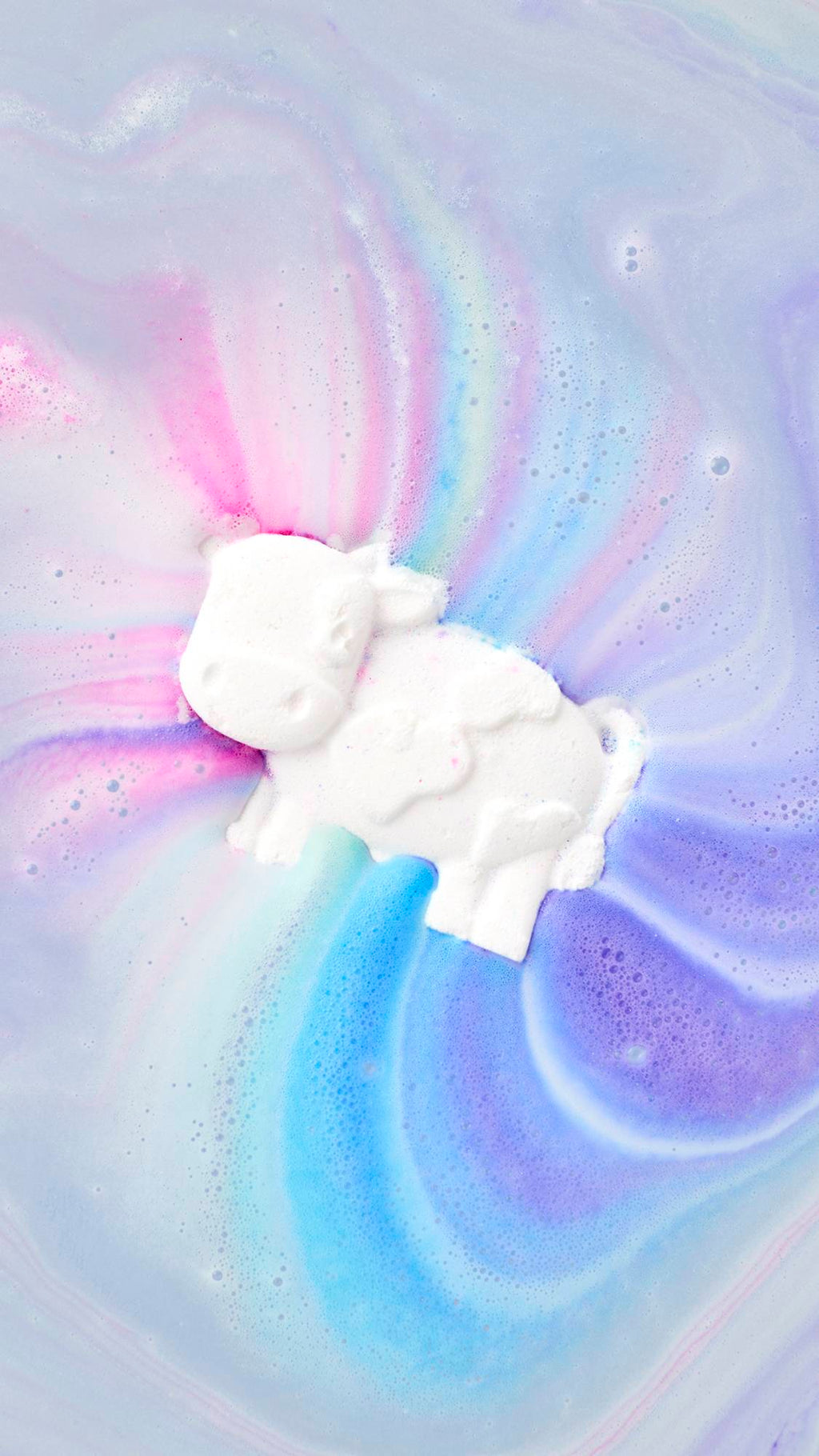 A top-down view of Toby's Magic Cow Bath Bomb as it dissolves in water, releasing a rainbow of colors. The white, cow-shaped bath bomb floats in the center, creating swirls of bright pink, blue, and purple fanning out across pastel purple bathwater.