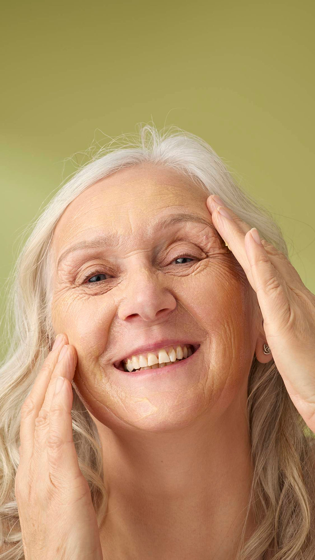 A model with long grey hair smiles while applying the yellow Turmeric And Argan cleanser to her face. She is shown from the shoulders up against a solid olive-green background, highlighting the creamy texture of the facial product.