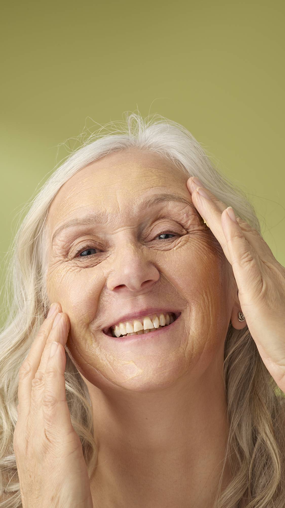 A model with long grey hair smiles while applying the yellow Turmeric And Argan cleanser to her face. She is shown from the shoulders up against a solid olive-green background, highlighting the creamy texture of the facial product.