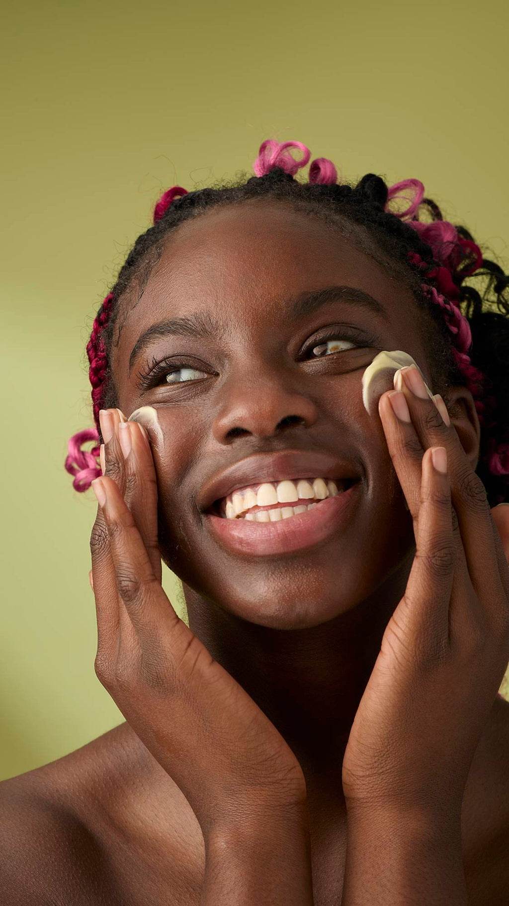 A person with pink braids smiles broadly while massaging Ultrabland cleanser onto their cheeks with both hands, looking upward against an olive green background.