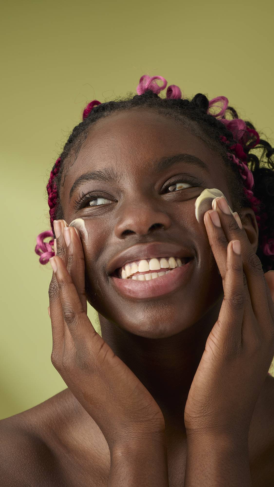 A person with pink braids smiles broadly while massaging Ultrabland cleanser onto their cheeks with both hands, looking upward against an olive green background.