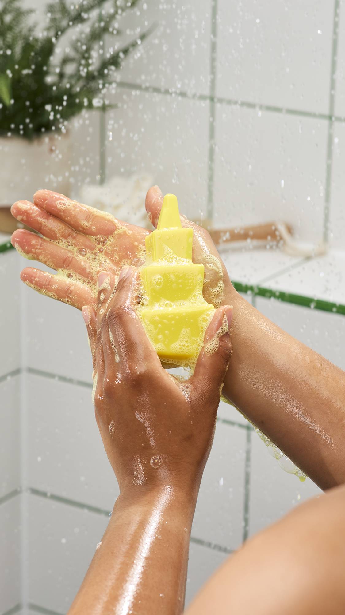Hands lathering a bright yellow, tiered geometric shower gummy soap under running water. Bubbles cover the hands against a background of white tiles with green grout and splashing water.