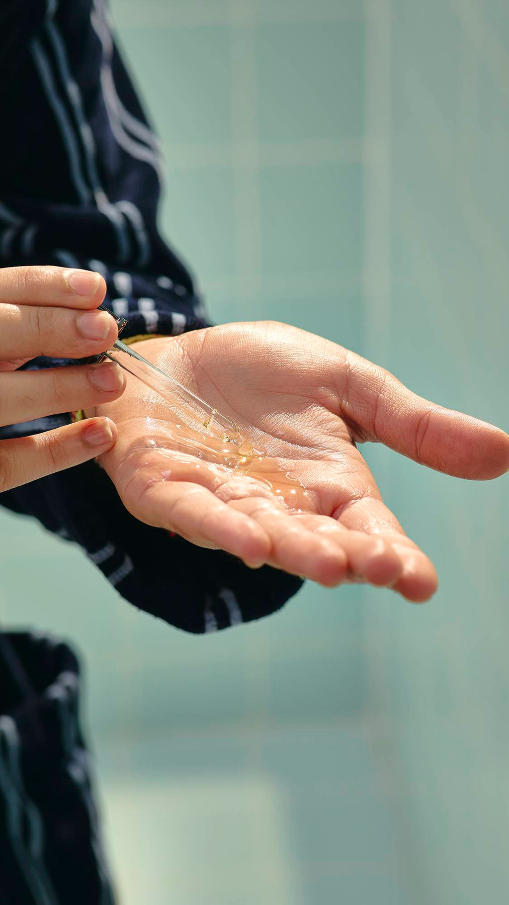 The image shows a close-up of a model's hands as they hold the glass bottle of "Vegan Leather Jacket" hair and beard oil and use the pipette topper to drop clear, golden oil into their open palm.