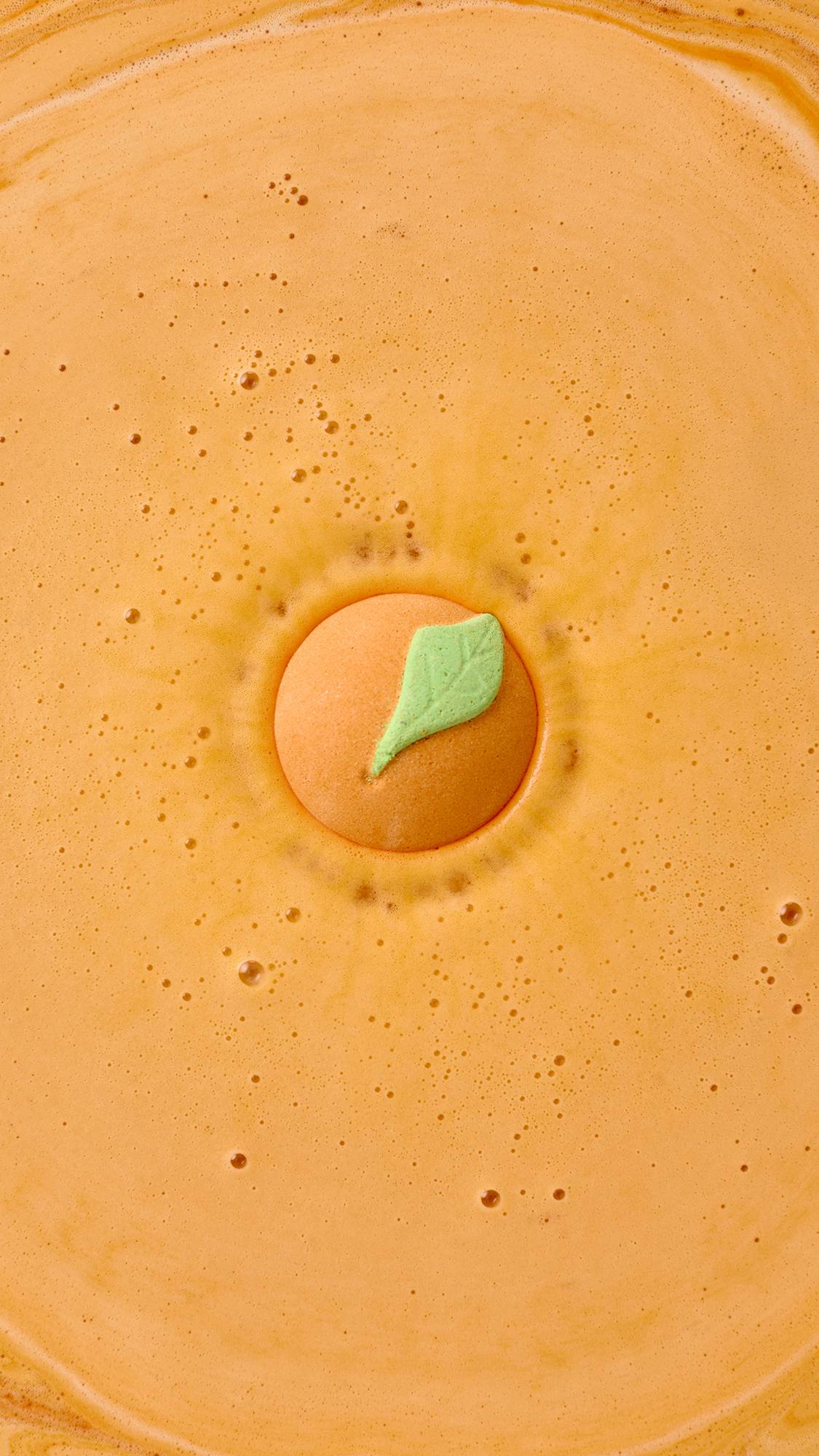 A top-down view shows the Peachy Bath Bomb dissolving in the center of a bathtub, turning the water a vibrant, opaque orange. The signature orange sphere, with its small green leaf, creates a bright and energizing soak.