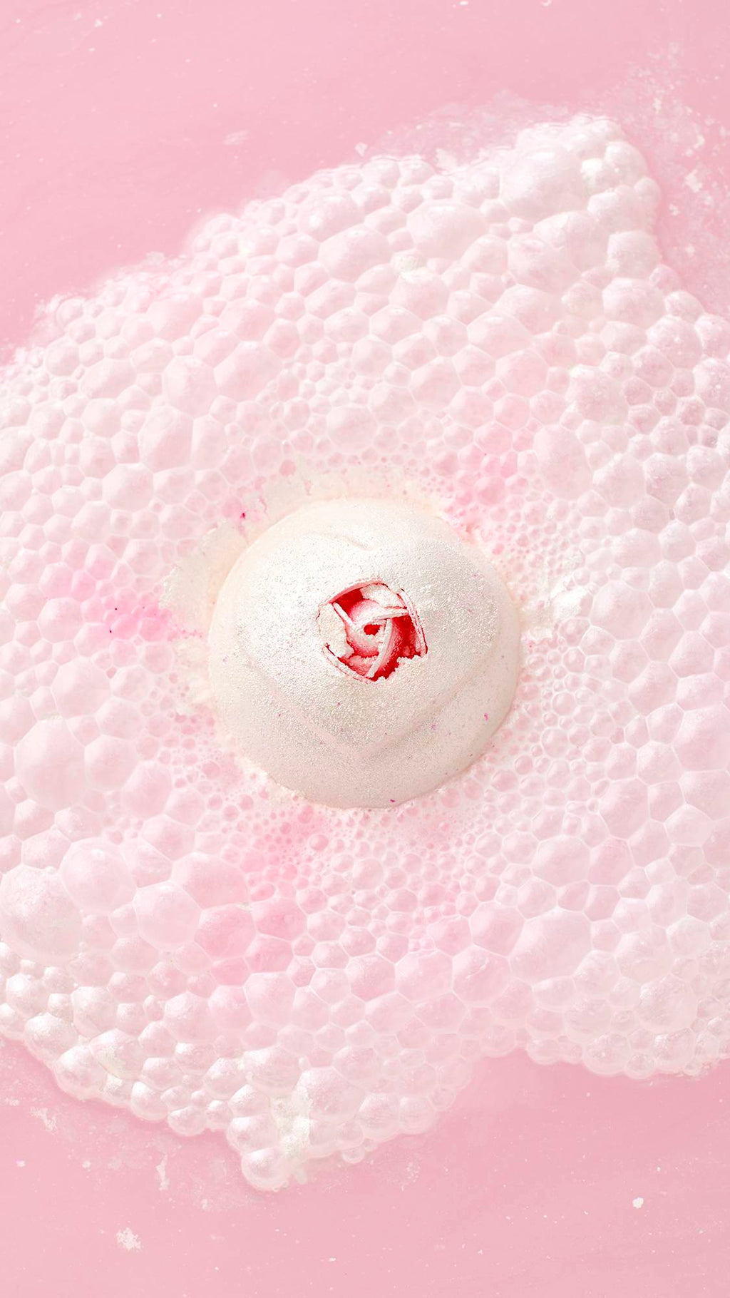 A top-down view of the white Wedding bath bomb with a pink rose center, floating in pink water. It generates a massive, textural cloud of white and pink bubbles and foam.