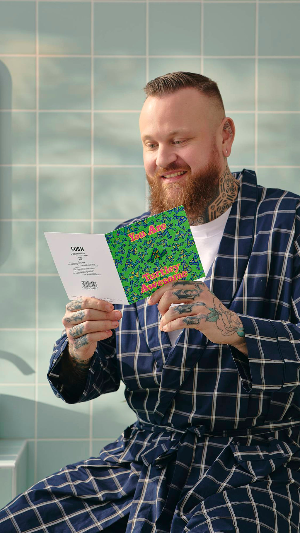 A model with a beard and tattoos, wearing a blue checked dressing gown, smiles while reading the inside of a 'Turtley Awesome' card, with a removable enamel turtle pin badge. They are in a bathroom with pale blue tiles in the background.