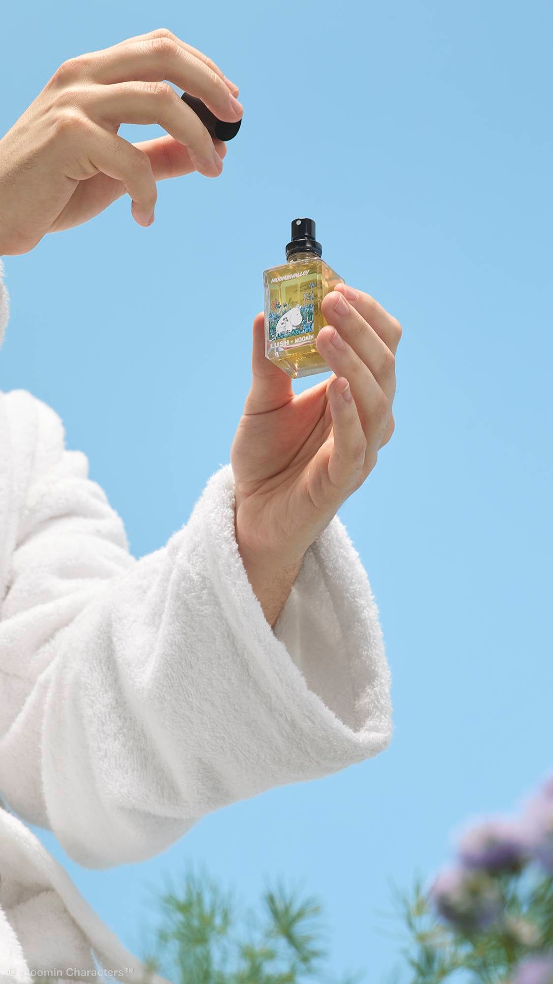 A close-up shot of a person's hands in a white, fluffy dressing gown holding up a small glass bottle of Lush x Moomin perfume against a clear blue sky. One hand gently holds the bottle whilst the other lifts off the black lid.