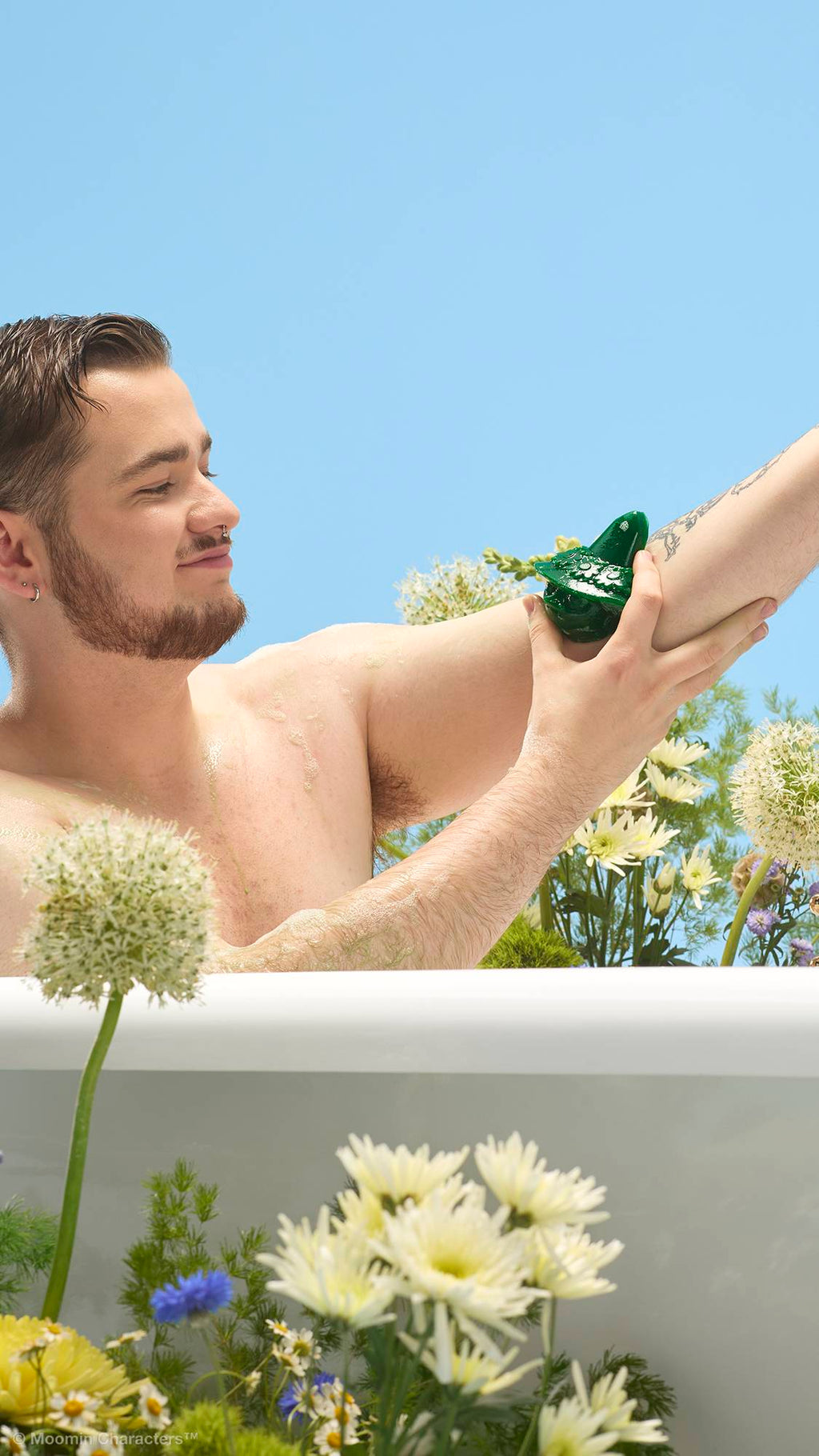 A close-up shot of a person with a beard sitting in a white bathtub, washing their arm with a green Snufkin-shaped shower gummy. Yellow and white wildflowers surround the tub against a clear blue sky background.