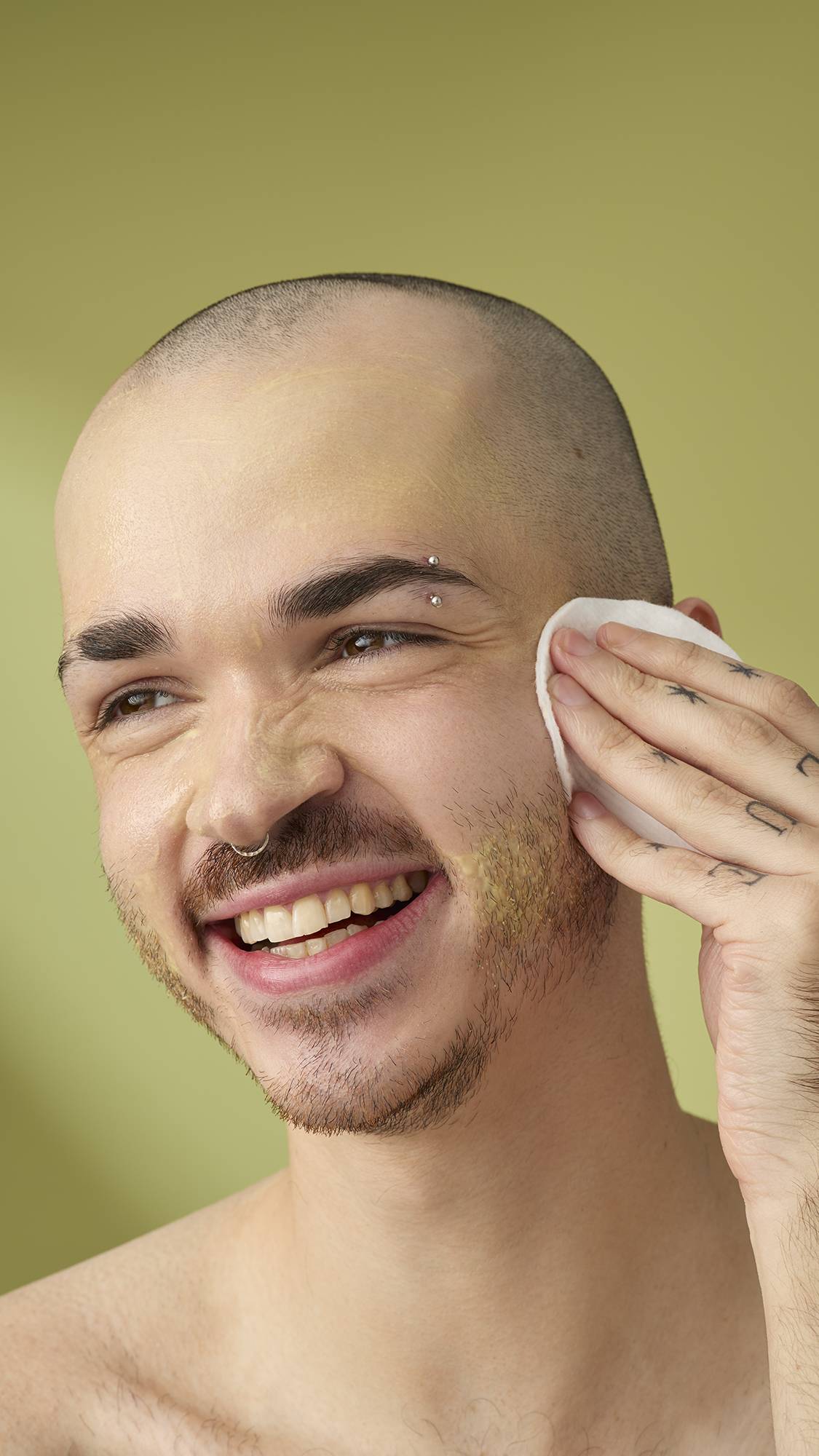 A person with a shaved head and facial piercings smiles while using a white cotton pad to wipe the Acai And Sacha Inchi cleanser from their cheek. They have tattooed hands and are shown against a solid olive-green background.