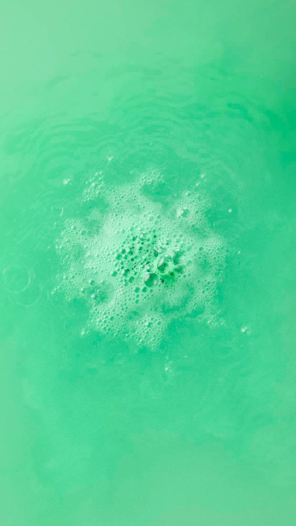 Top-down view of the Avobath Bath bomb dissolving in vibrant green water. A concentrated cluster of frothy, light green bubbles fizzes in the center of the shimmering, lime-colored bathwater.