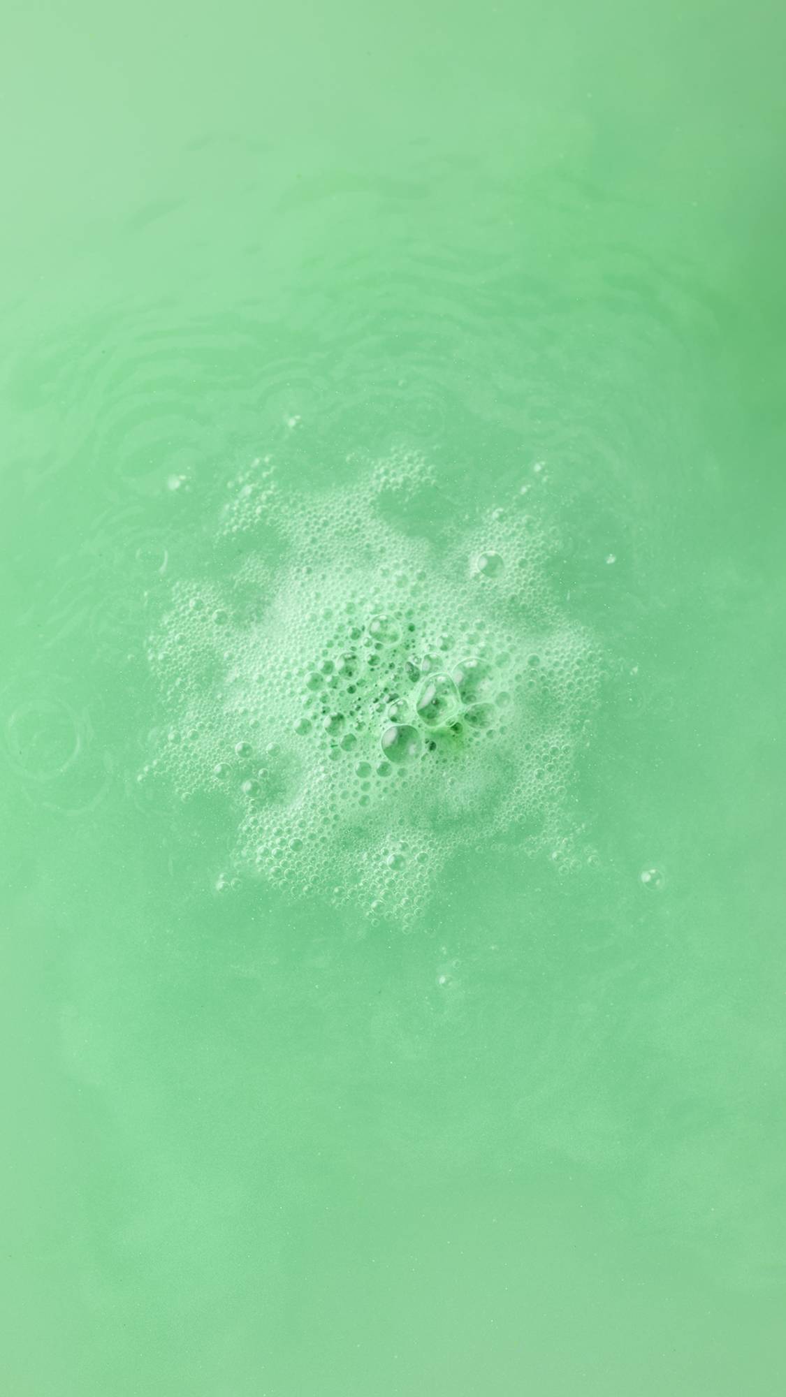 Top-down view of the Avobath Bath bomb dissolving in vibrant green water. A concentrated cluster of frothy, light green bubbles fizzes in the center of the shimmering, lime-colored bathwater.