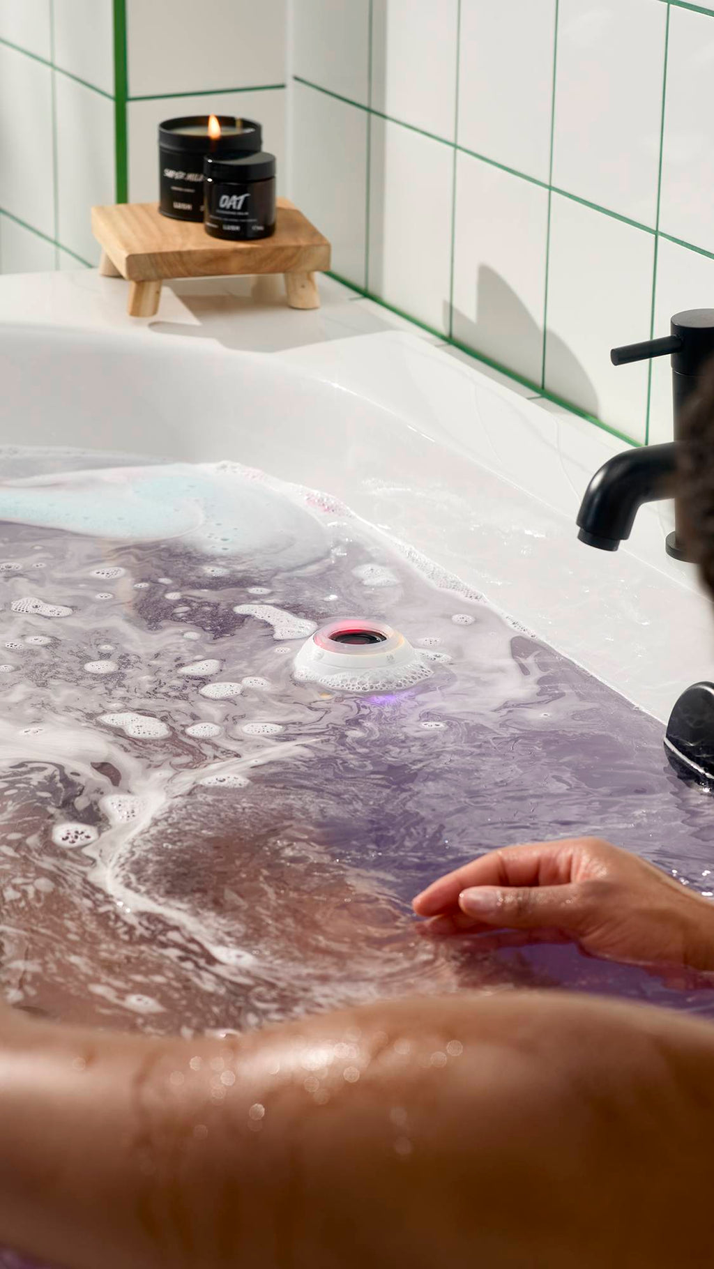 A person relaxes in a white bathtub filled with swirling purple water and white foam. A white bath bomb-shaped waterproof speaker with a red and blue glow floats in the centre. Lush candles and skin care sit on a wooden stand in the background.