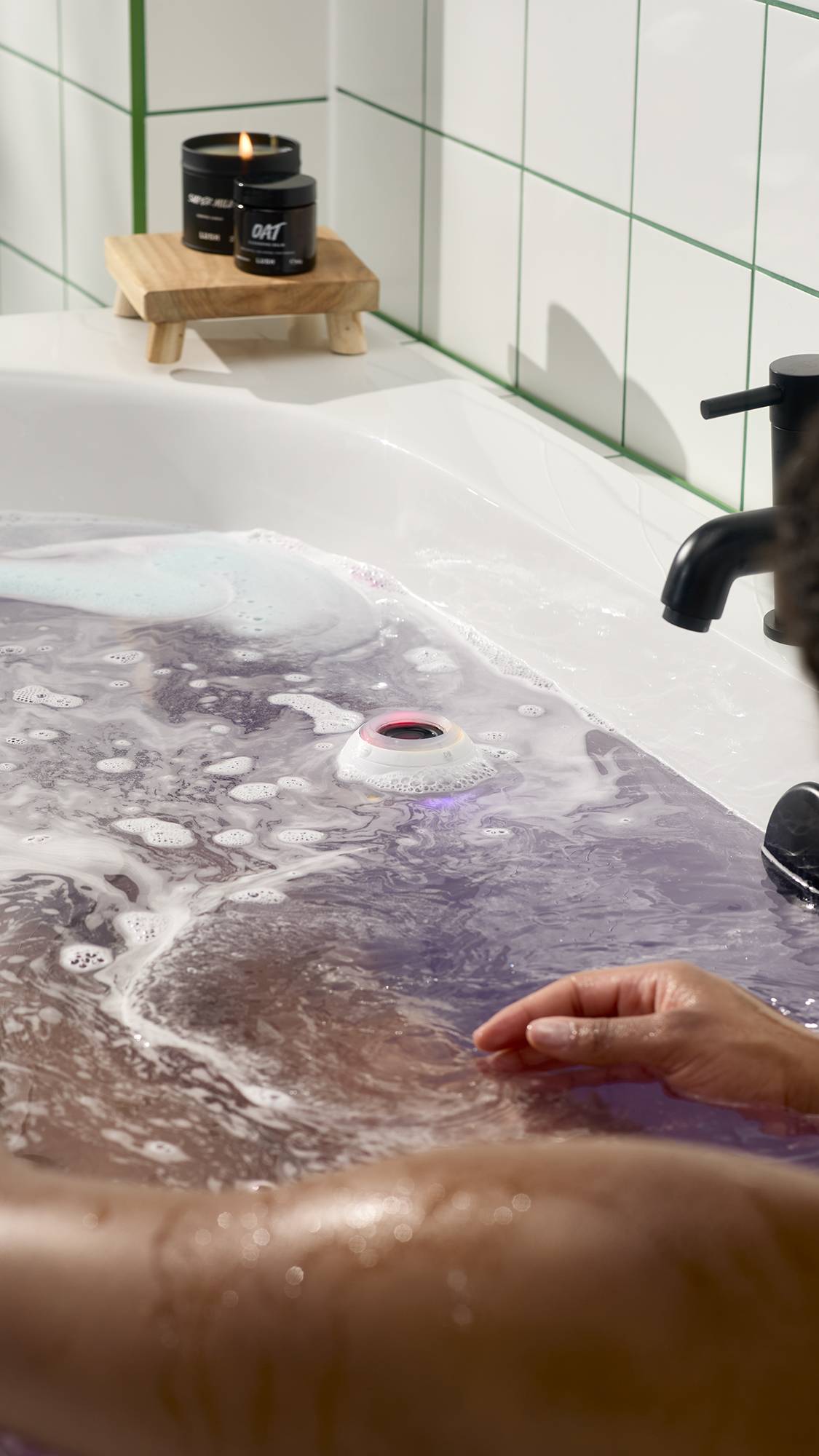 A person relaxes in a white bathtub filled with swirling purple water and white foam. A white bath bomb-shaped waterproof speaker with a red and blue glow floats in the centre. Lush candles and skin care sit on a wooden stand in the background.