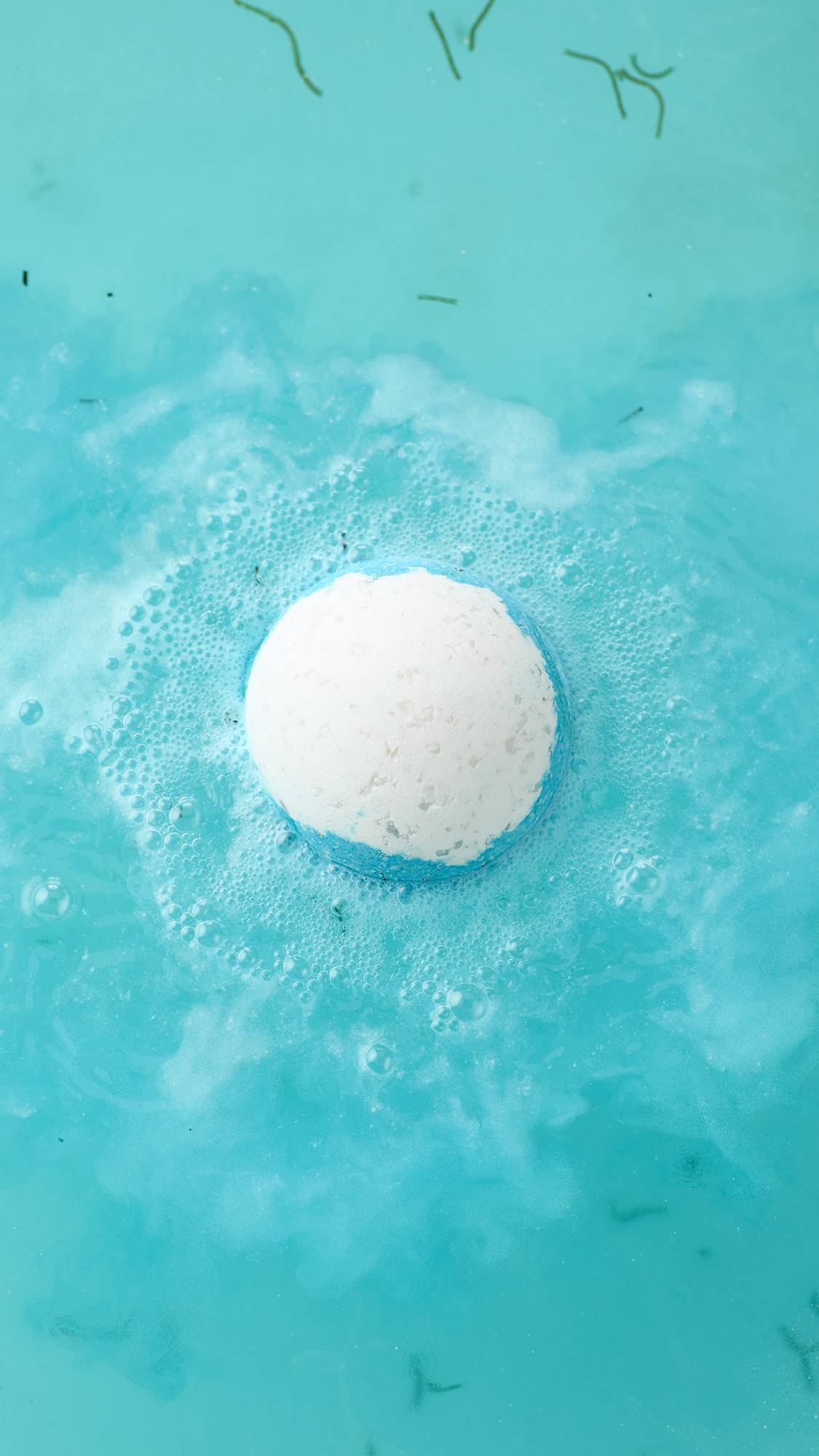 A top-down view of the Big Blue bath bomb fizzing in turquoise water. The white sphere is surrounded by a halo of blue and white foam, with small green seaweed strands floating in the vibrant bathwater.