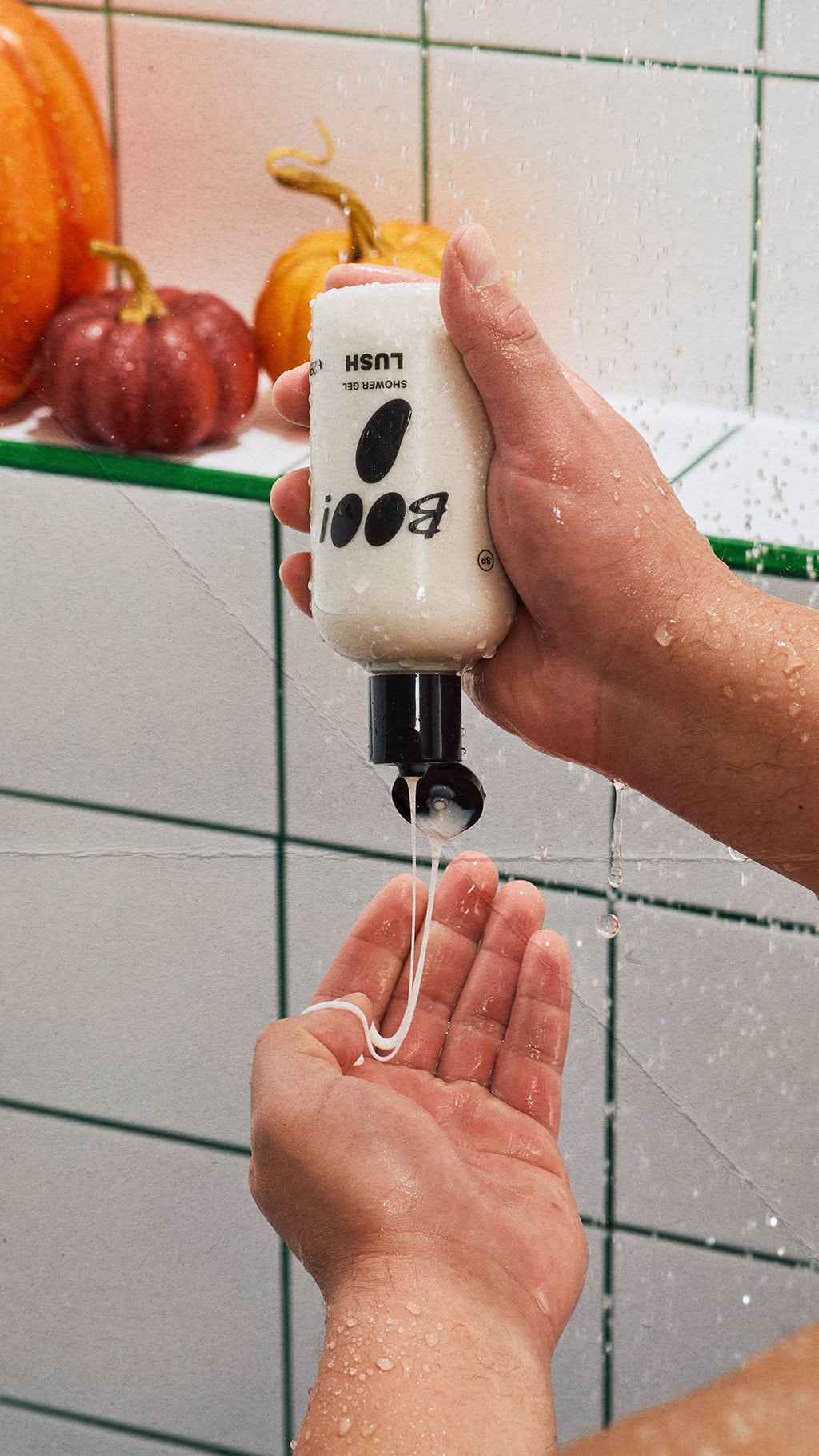 A person squeezes a bottle of Lush 'BOO!' shower gel upside down under running water, pouring the product into their other hand. White tiles with green grout form the background, with three decorative pumpkins visible on a ledge.