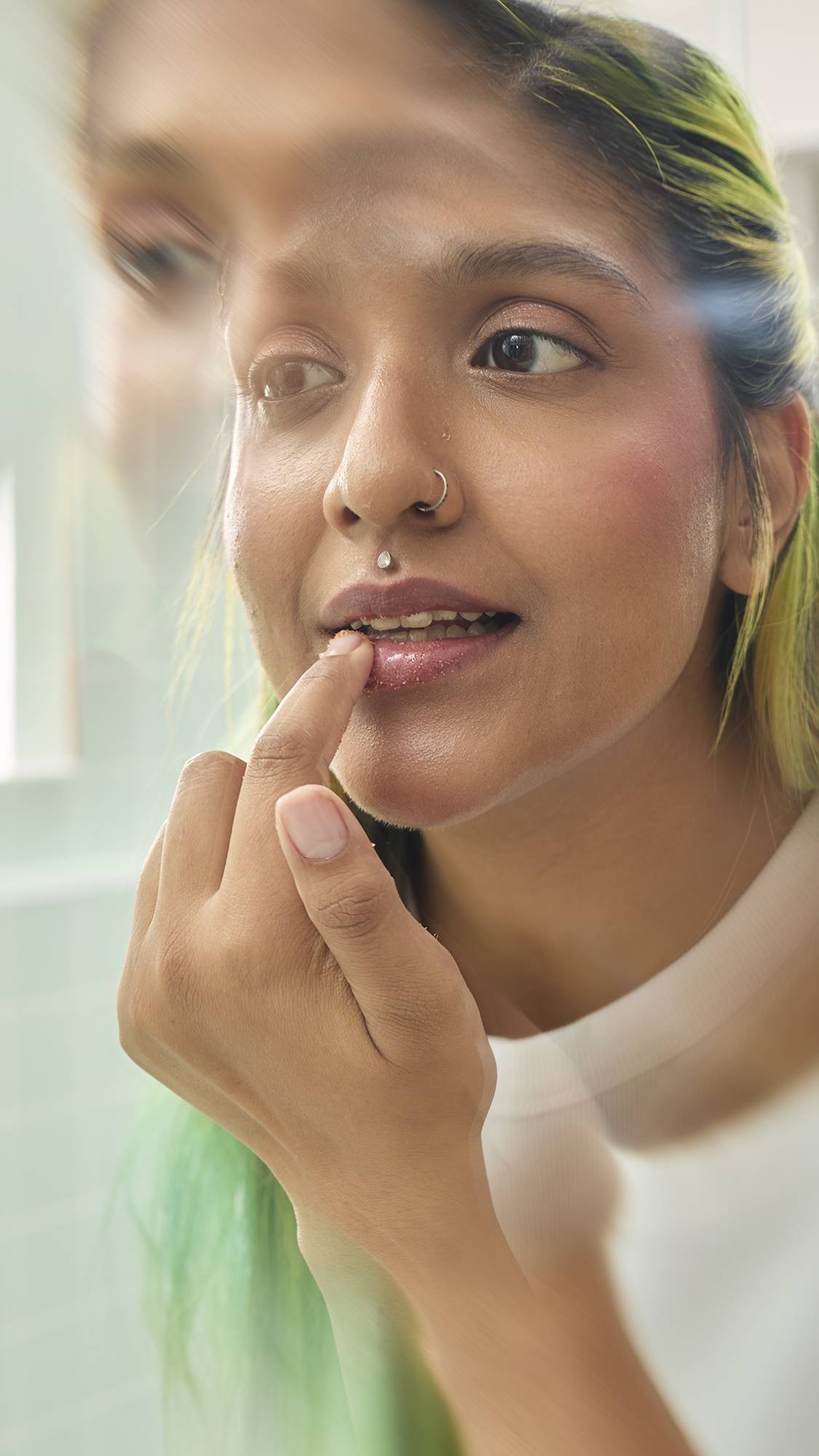 A close-up shot shows a model applying Lush "Buzz" Lip Scrub to their lips using their index finger. They have long, lime-green hair and facial piercings. The image is taken in a brightly lit bathroom, with a soft blur around the edges.